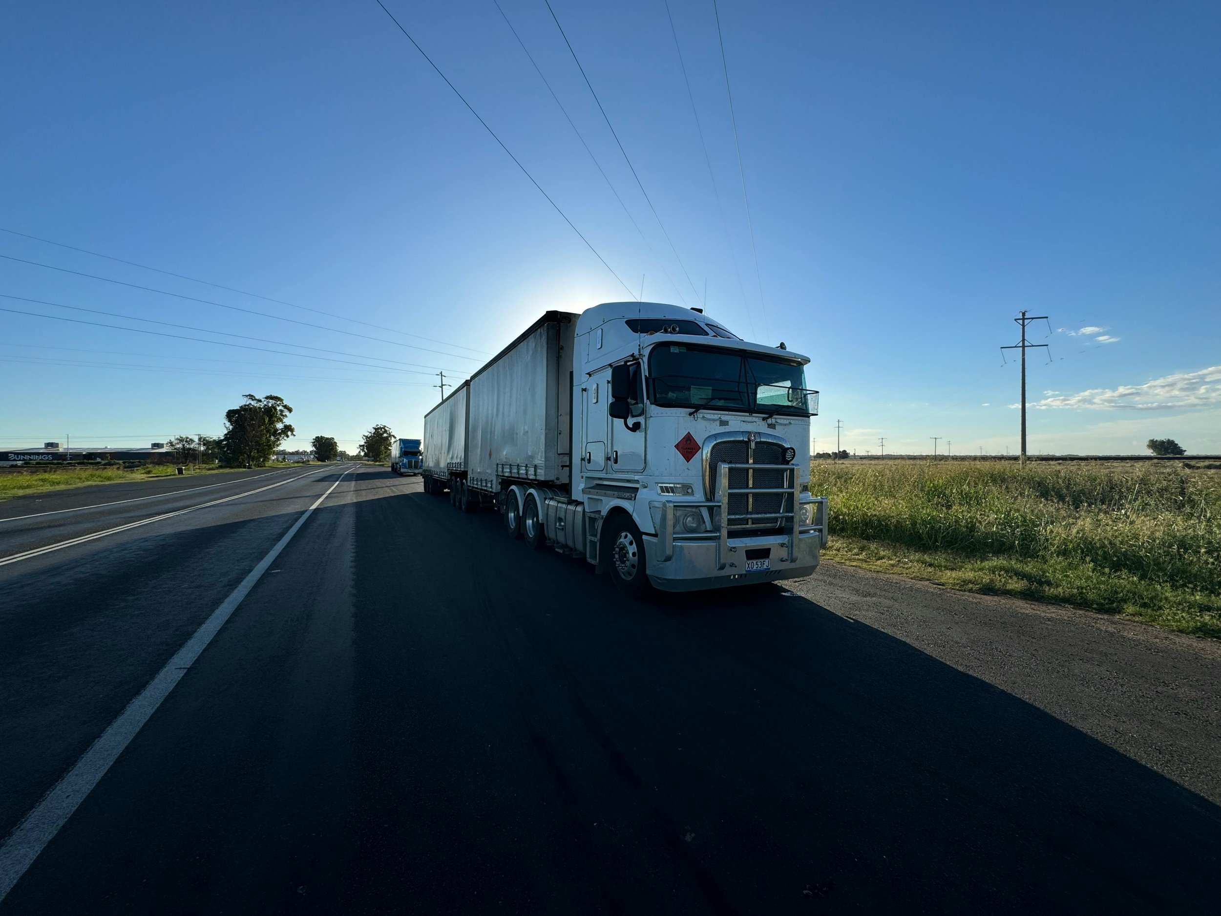 A white semi-truck parked on the shoulder of a rural highway with a clear blue sky and power lines in the background.