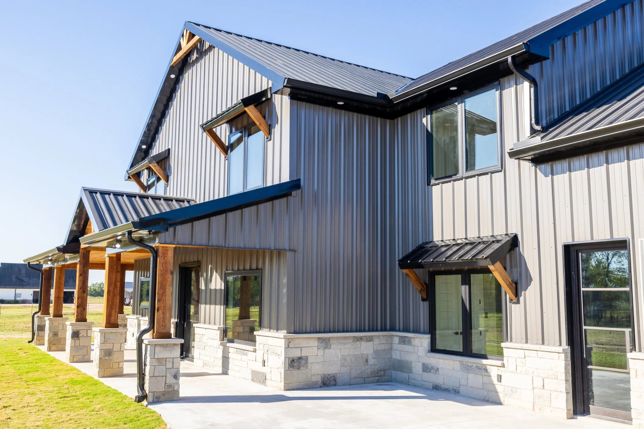 A modern two-story house with metal siding, multiple windows with small awnings, and a combination of wood and stone accents, surrounded by a neatly maintained lawn.