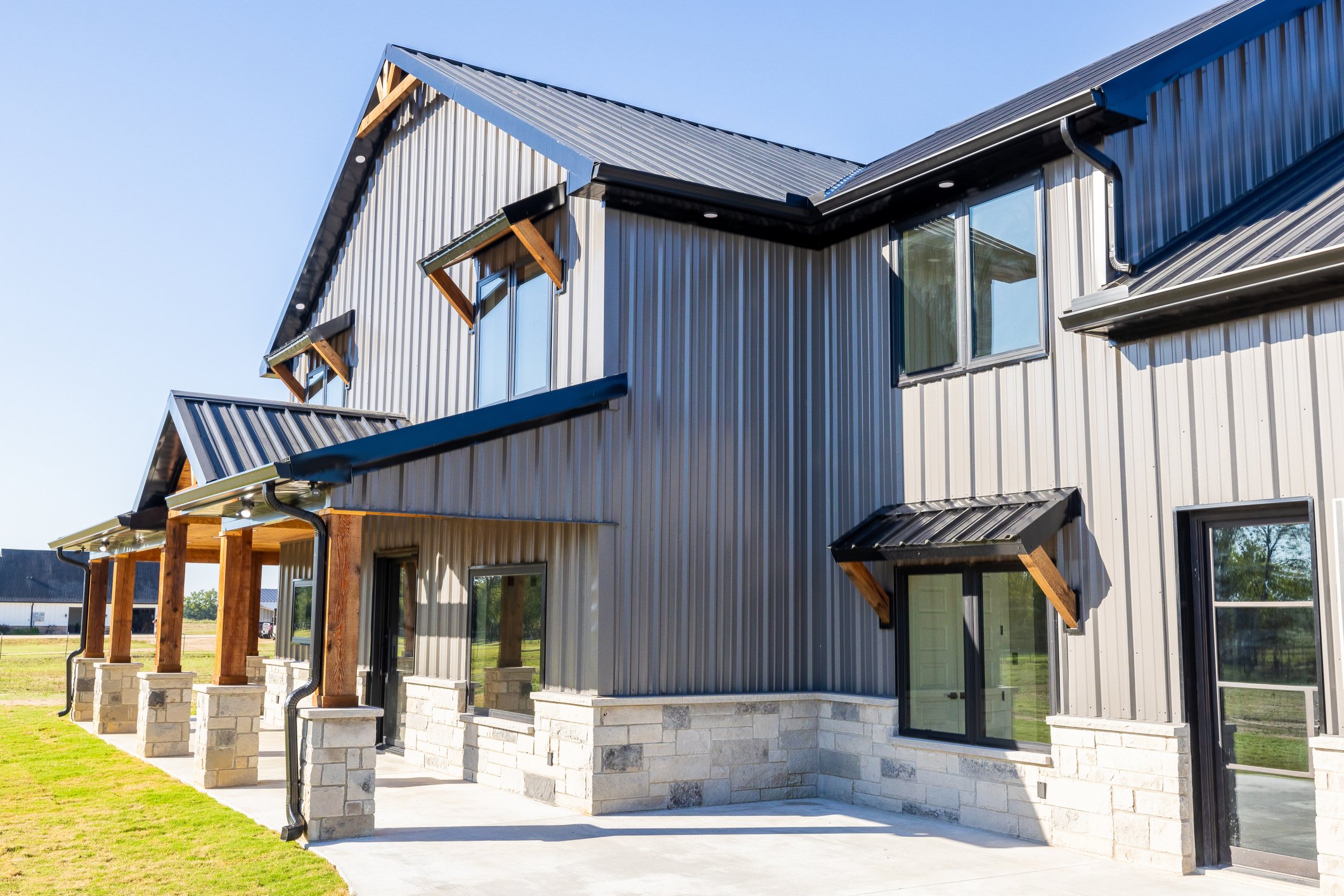 Modern building with metal siding, multiple windows with small awnings, and a concrete porch with brick supports, set against a clear blue sky.