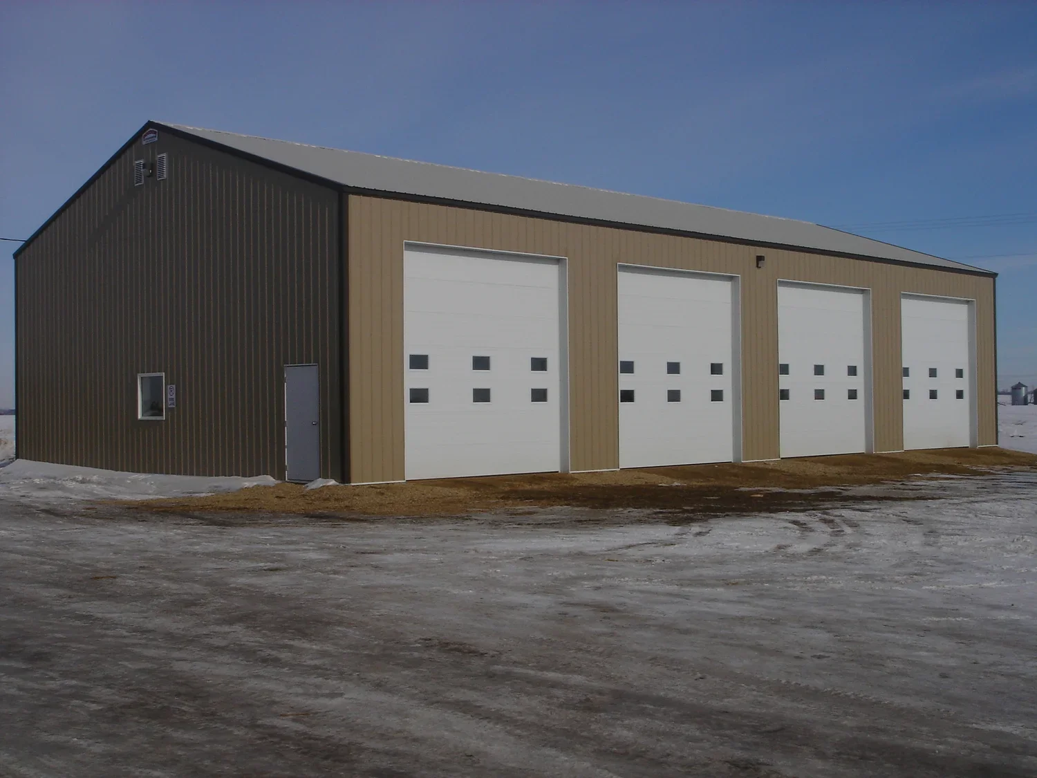 A large metal building with four white garage doors and a small side door, situated in a snow-covered landscape.