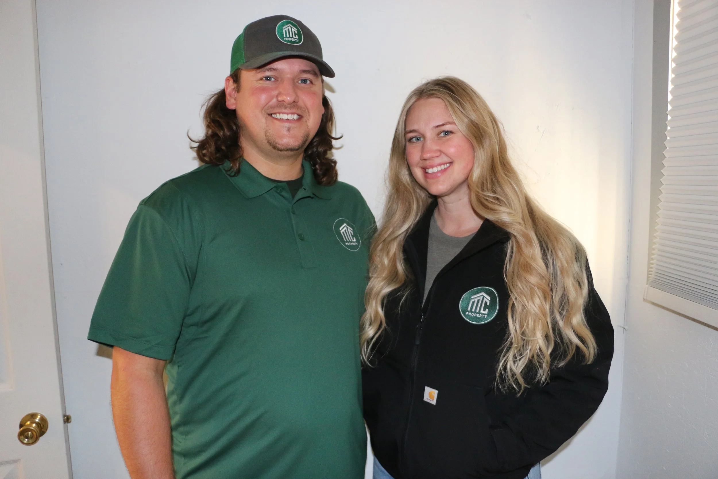A man and woman smiling, wearing work uniforms with a logo that says 'MC Property.' They are standing indoors against a white wall, with the man in a green polo and cap, and the woman in a black jacket.