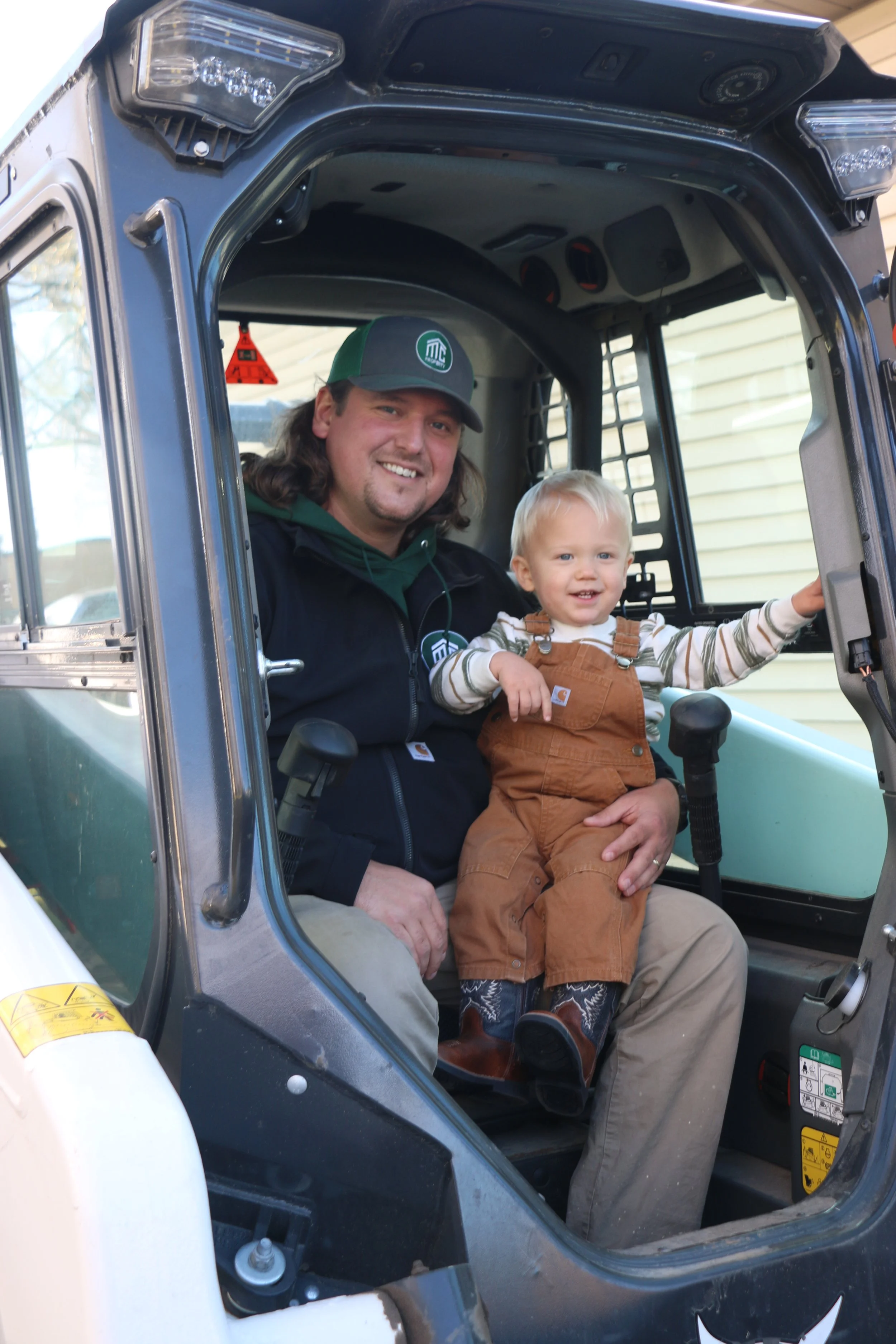 Man and young child sitting inside the cabin of a tractor, smiling at the camera.