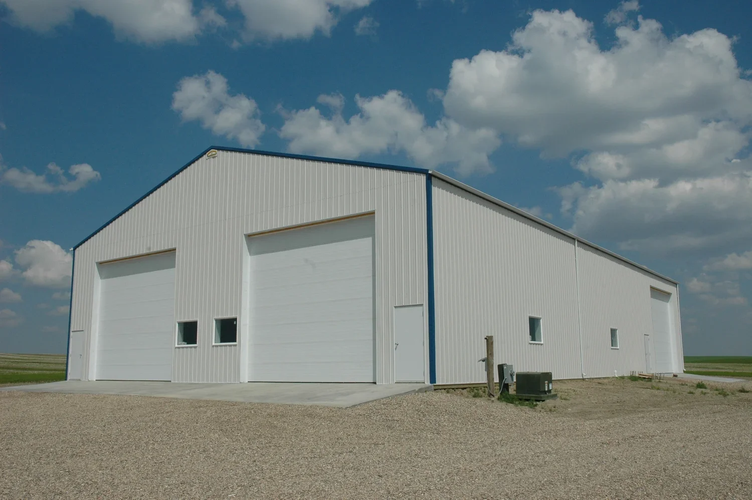 A large white metal storage building with two garage doors, small windows, and a gravel driveway under a partly cloudy sky.