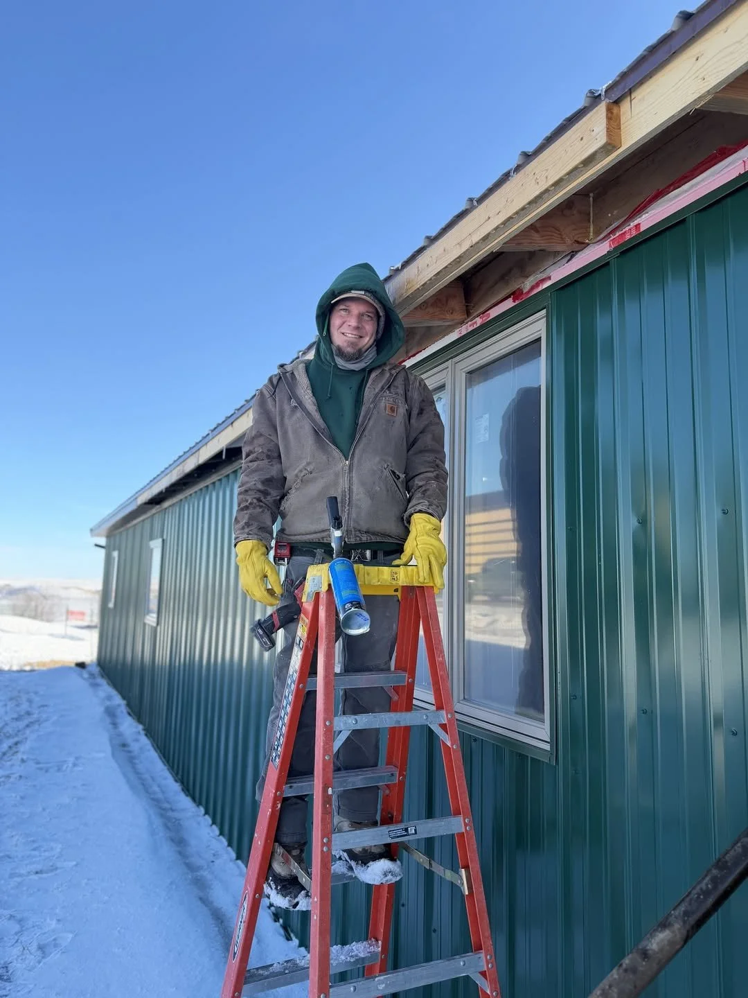 A man standing on a red ladder outside a green metal building under a clear blue sky, holding a caulking gun and wearing yellow gloves and warm clothing, including a hoodie and a jacket, during winter.