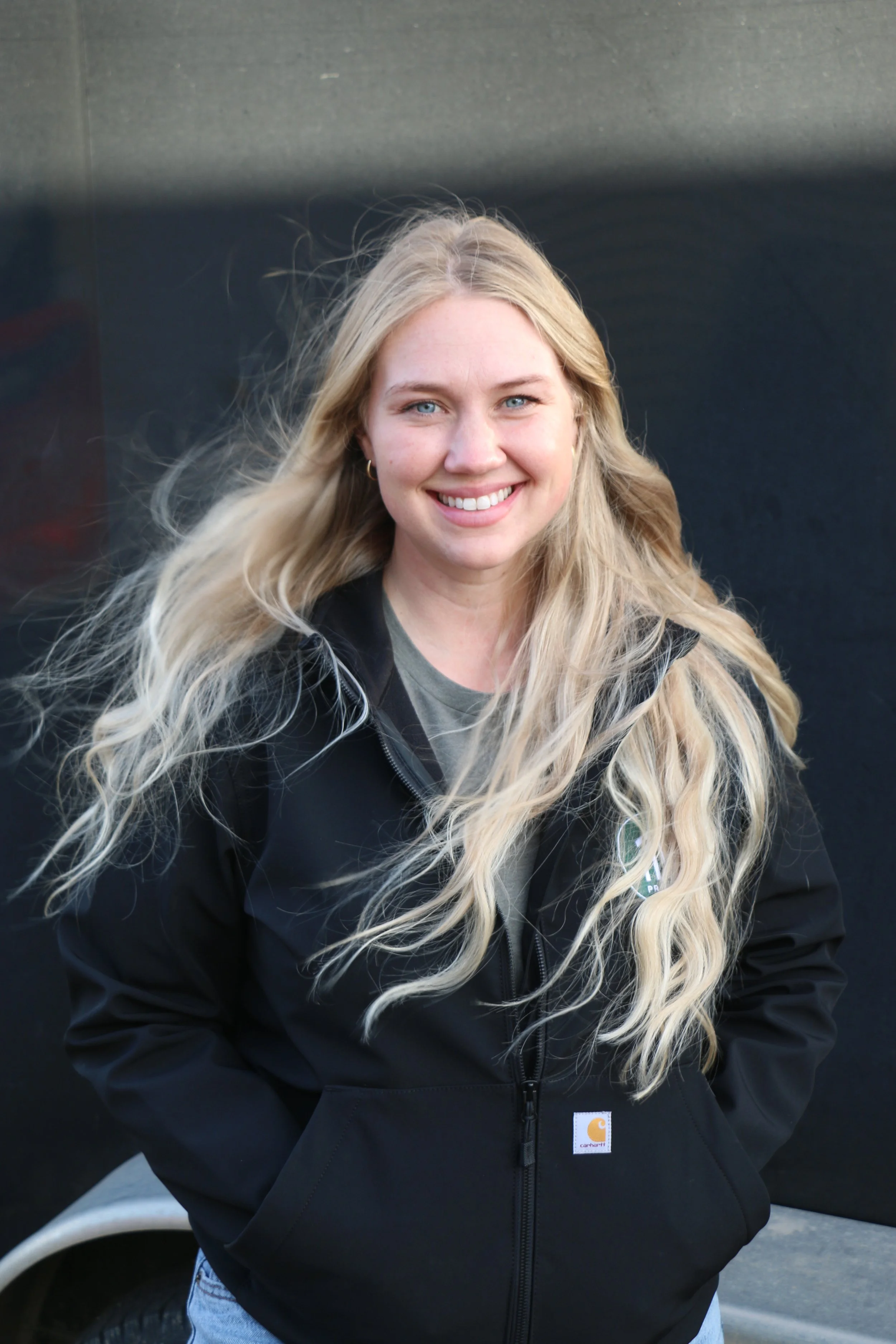 A young woman with long, wavy blonde hair and blue eyes, smiling while wearing a black jacket over a gray shirt, standing outdoors against a dark background.