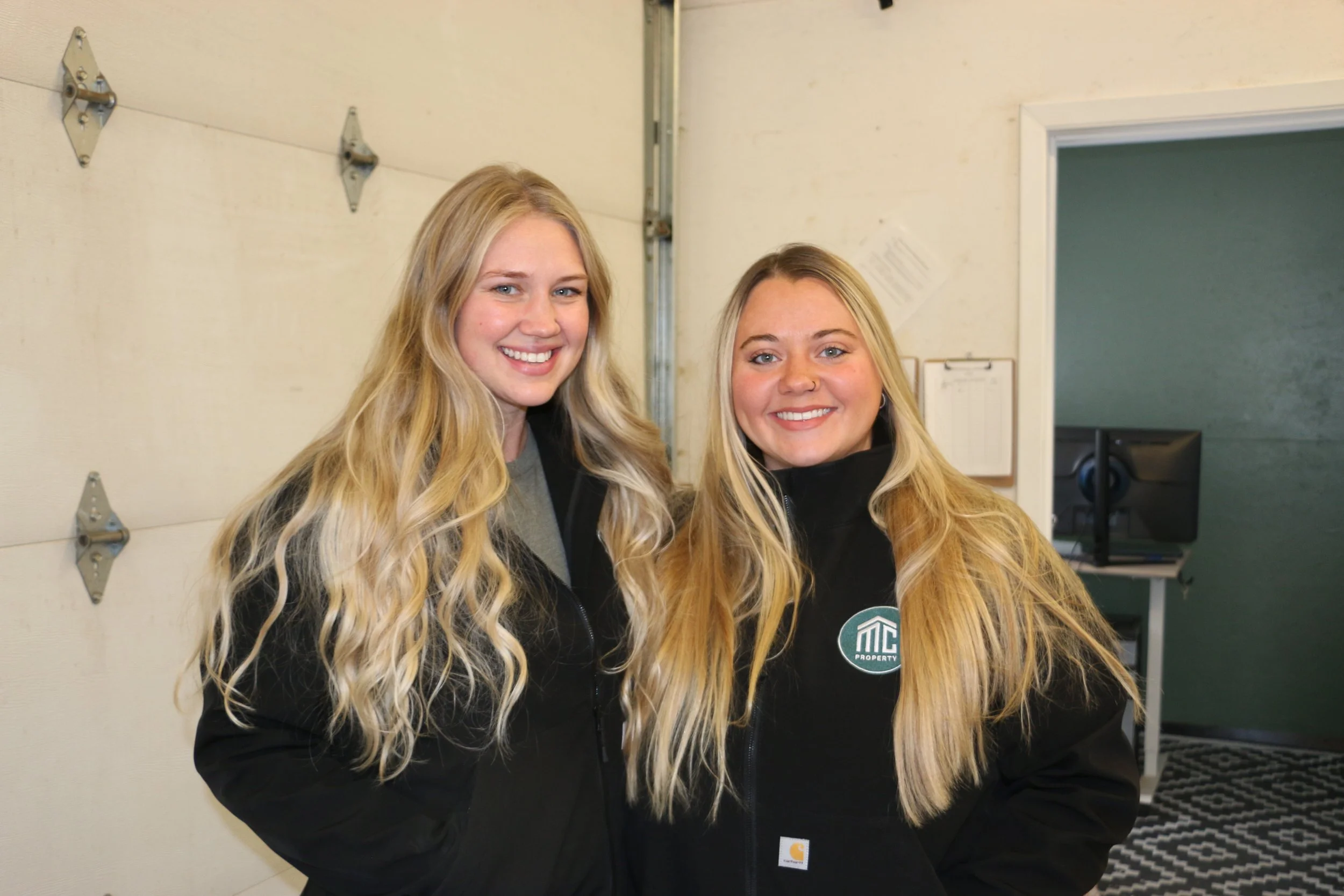 Two smiling young women with long blonde hair standing inside a room near a garage door and a green wall, both wearing black jackets.
