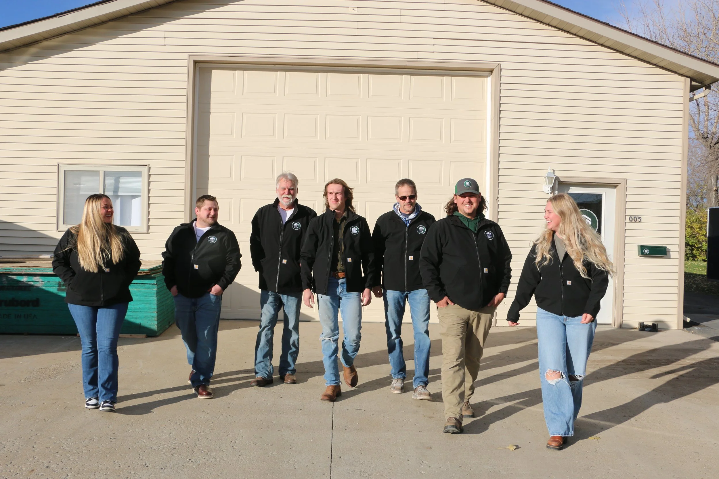 Group of eight people walking in front of a beige building with a large garage door, all wearing matching black jackets with a circular logo.