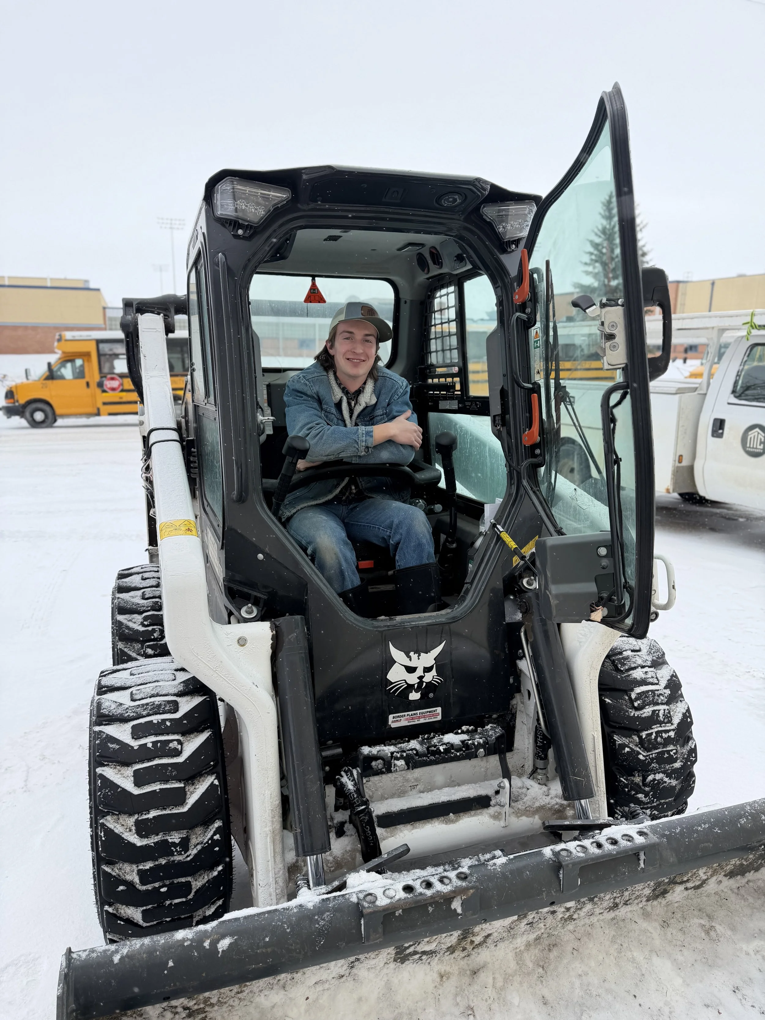 A young man with long brown hair smiling, standing outdoors near construction equipment, wearing a black jacket over a plaid shirt and jeans.