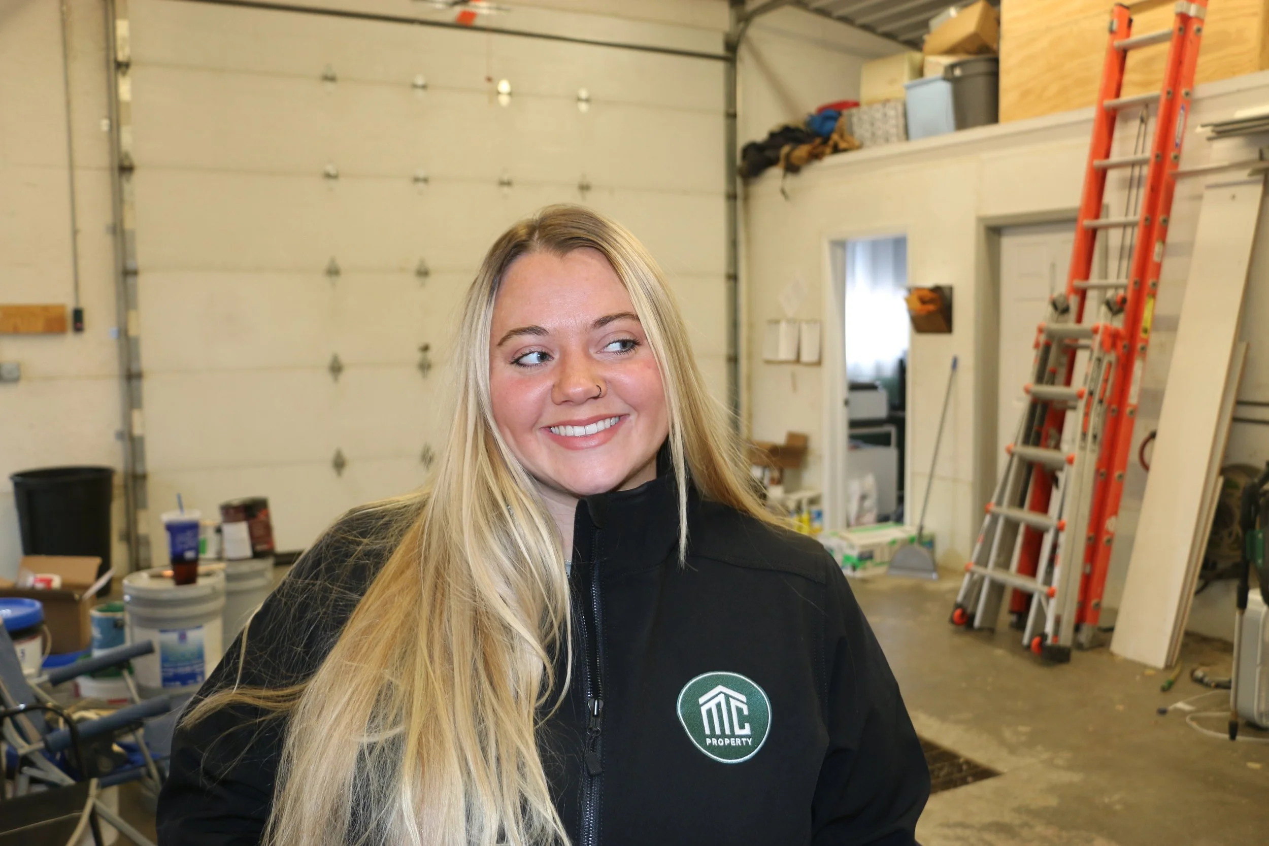 A smiling woman with long blonde hair, wearing a black jacket with a logo that says 'MC Property,' standing inside a garage or workshop.