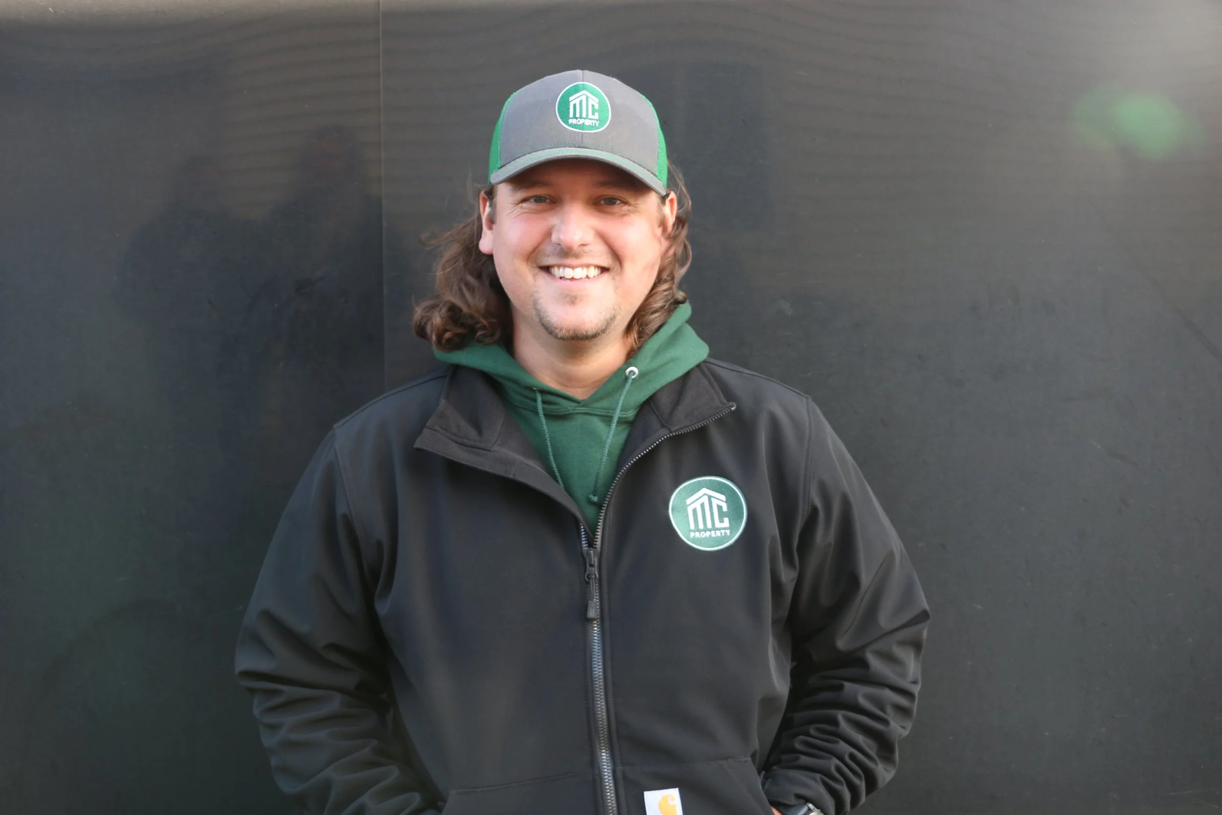 A smiling man with long brown hair wearing a black jacket, green hoodie, and a gray and green cap with a logo, standing in front of a dark background.