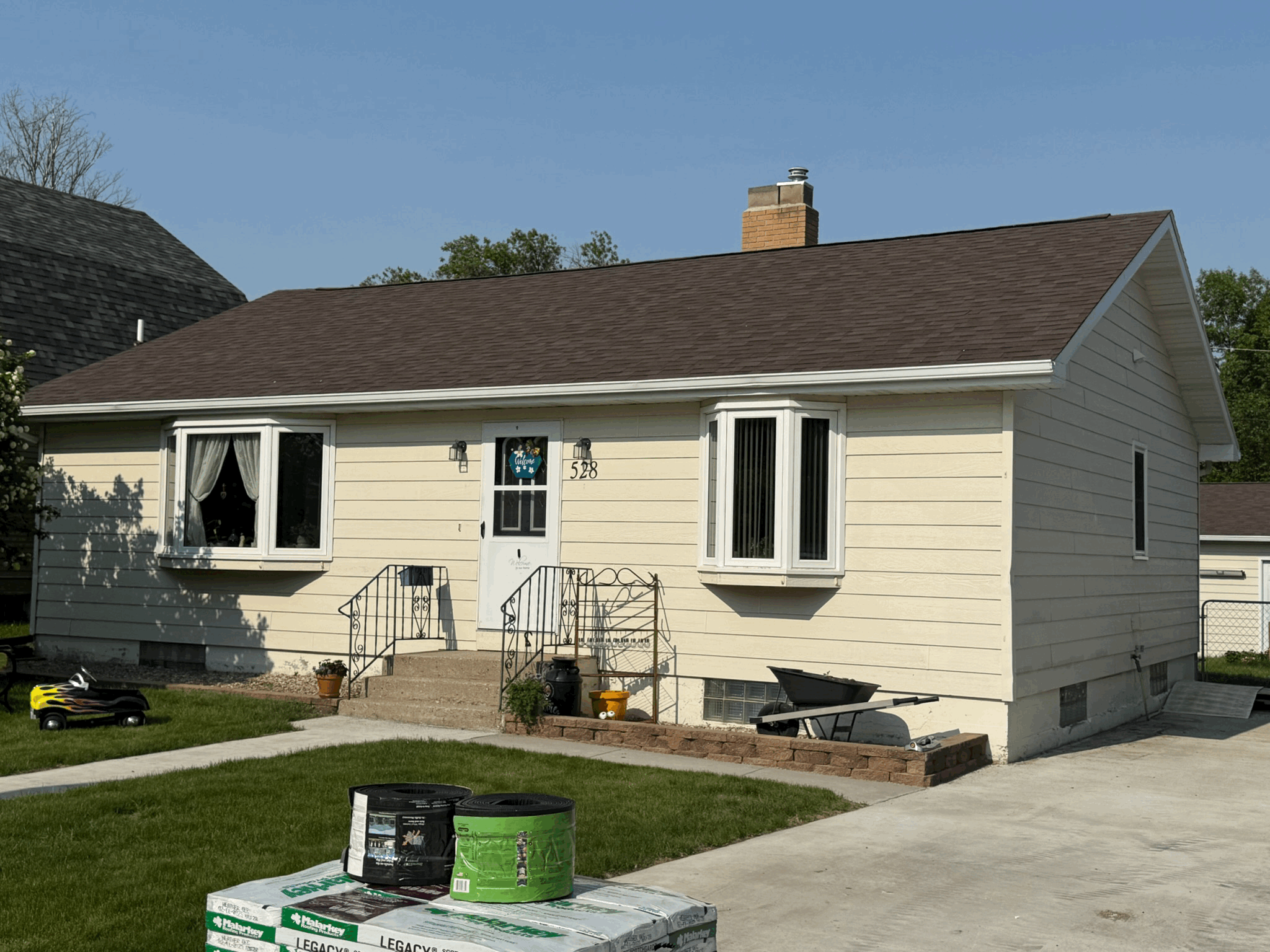 Front view of a suburban house with yellow siding, brown roof, and small steps leading to the front door, with patchy green lawn, and construction materials in the foreground.