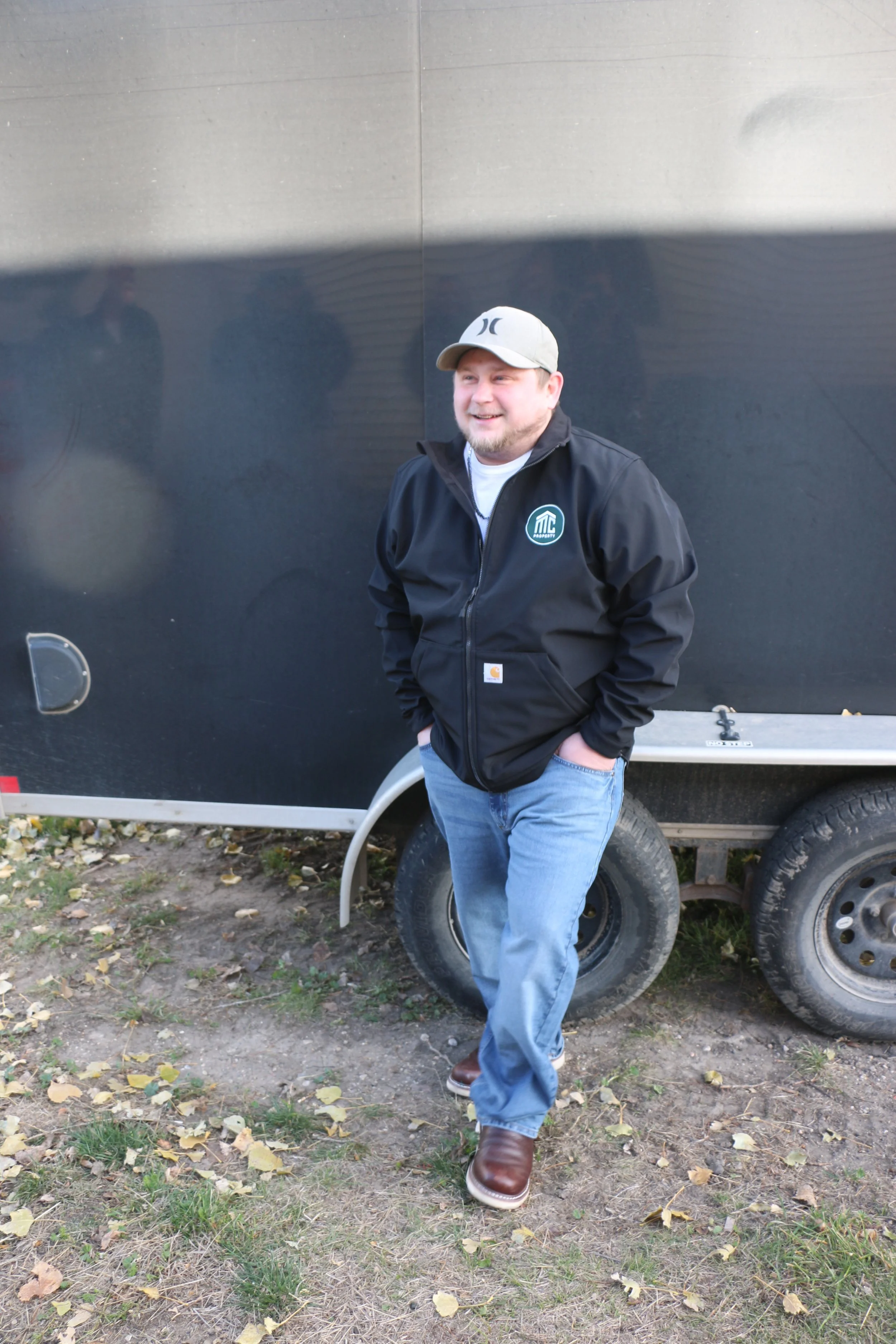 A man in a black jacket, blue jeans, and brown boots standing outdoors in front of a black trailer, smiling.