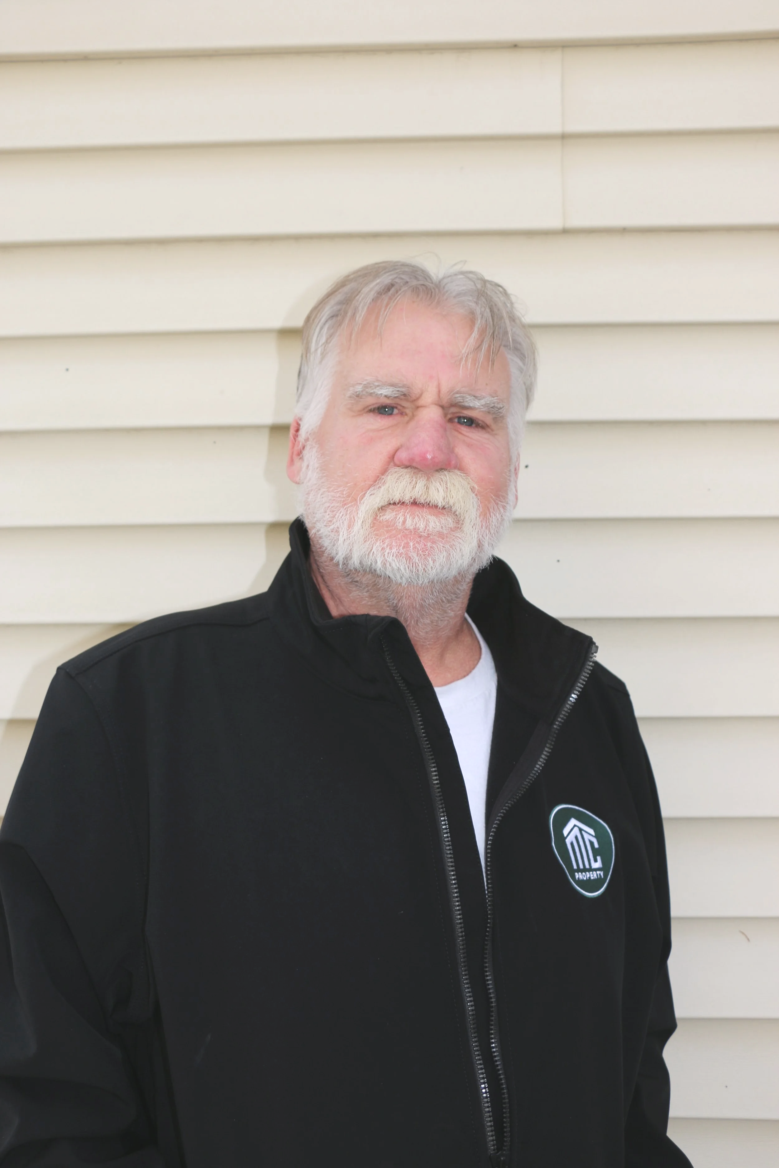 Older man with white hair and beard, wearing a black jacket with a logo, standing outside in front of beige horizontal siding.