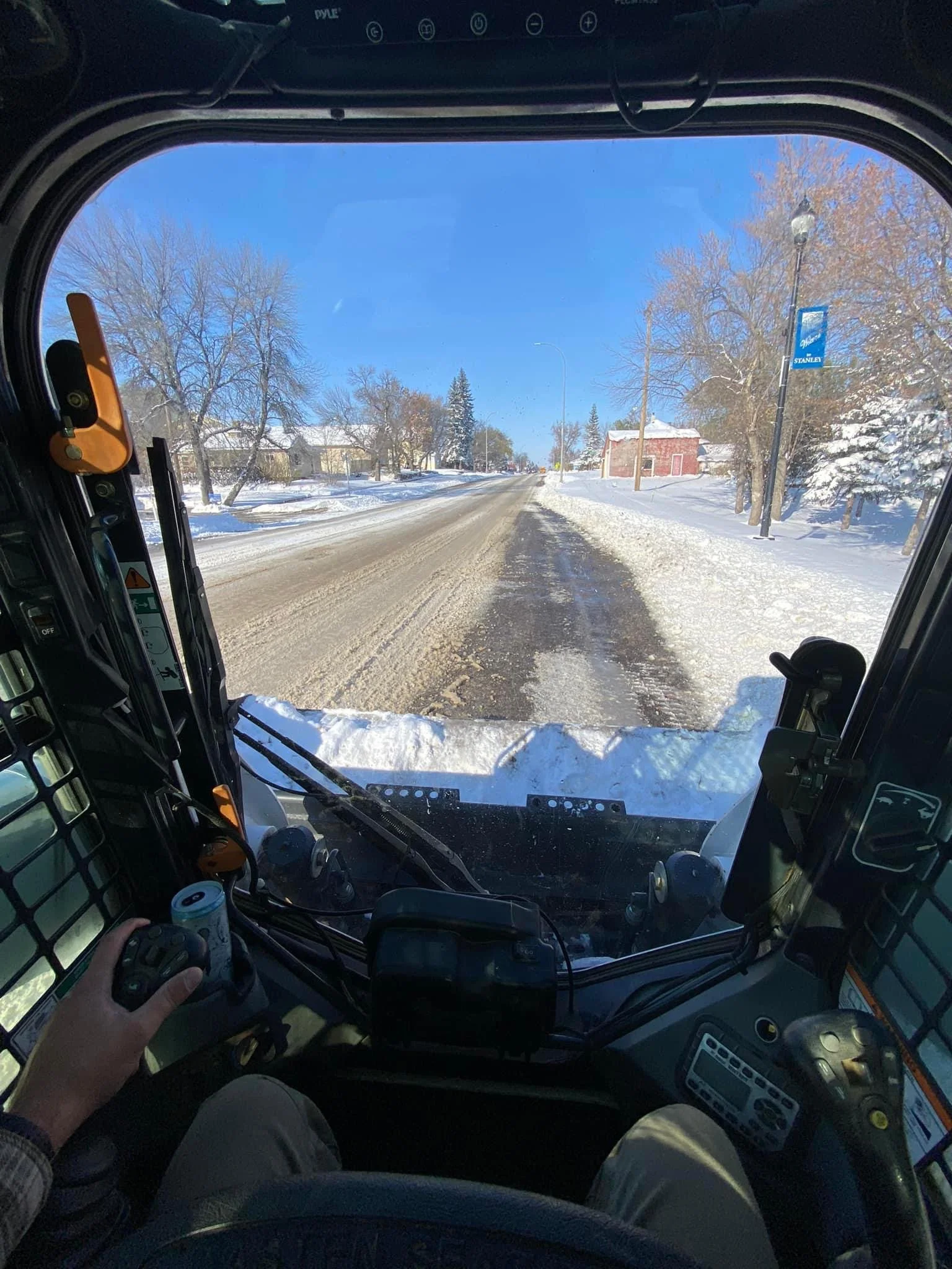 Inside the cab of a snow-covered tractor or construction vehicle, looking out over a snowy, icy road with buildings, trees, and a street lamp visible under a clear blue sky.