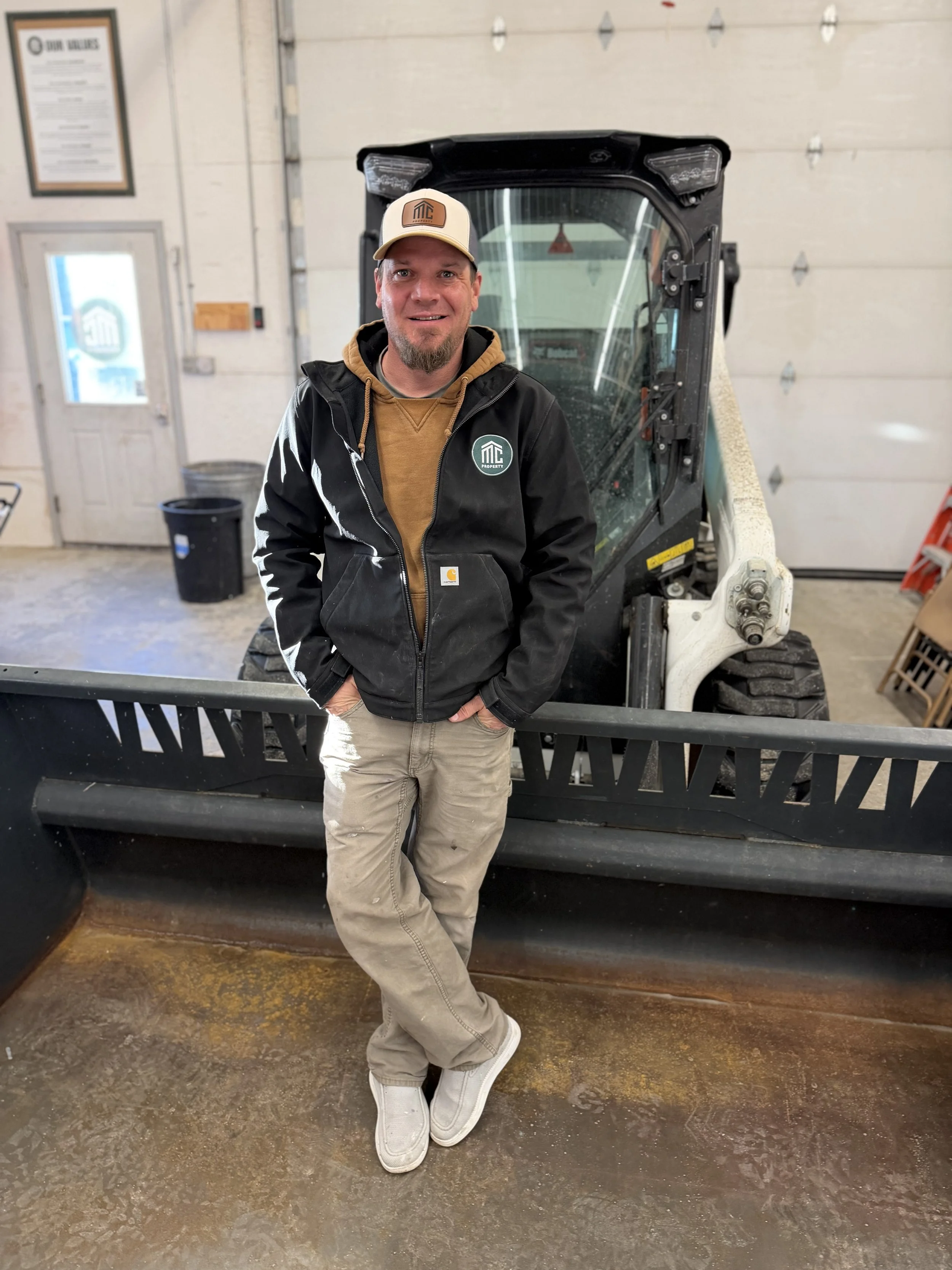 A man standing on a red ladder outside a green metal building under a clear blue sky, holding a caulking gun and wearing yellow gloves and warm clothing, including a hoodie and a jacket, during winter.