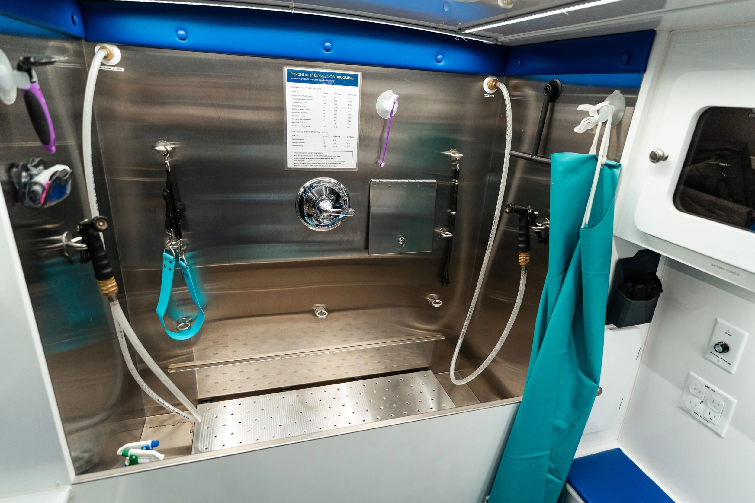 Interior of a mobile dog grooming station with stainless steel walls, hoses, grooming tools, and a foldable grooming table with a blue privacy curtain.