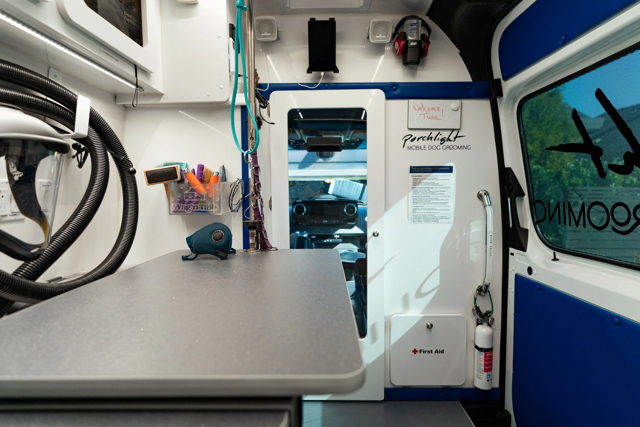 The interior of a mobile dog grooming van with a grooming station, storage shelves, and grooming supplies. There is a mirror, a whiteboard that says 'Welcome! Tuna', and equipment such as hoses, brushes, and a small speaker.