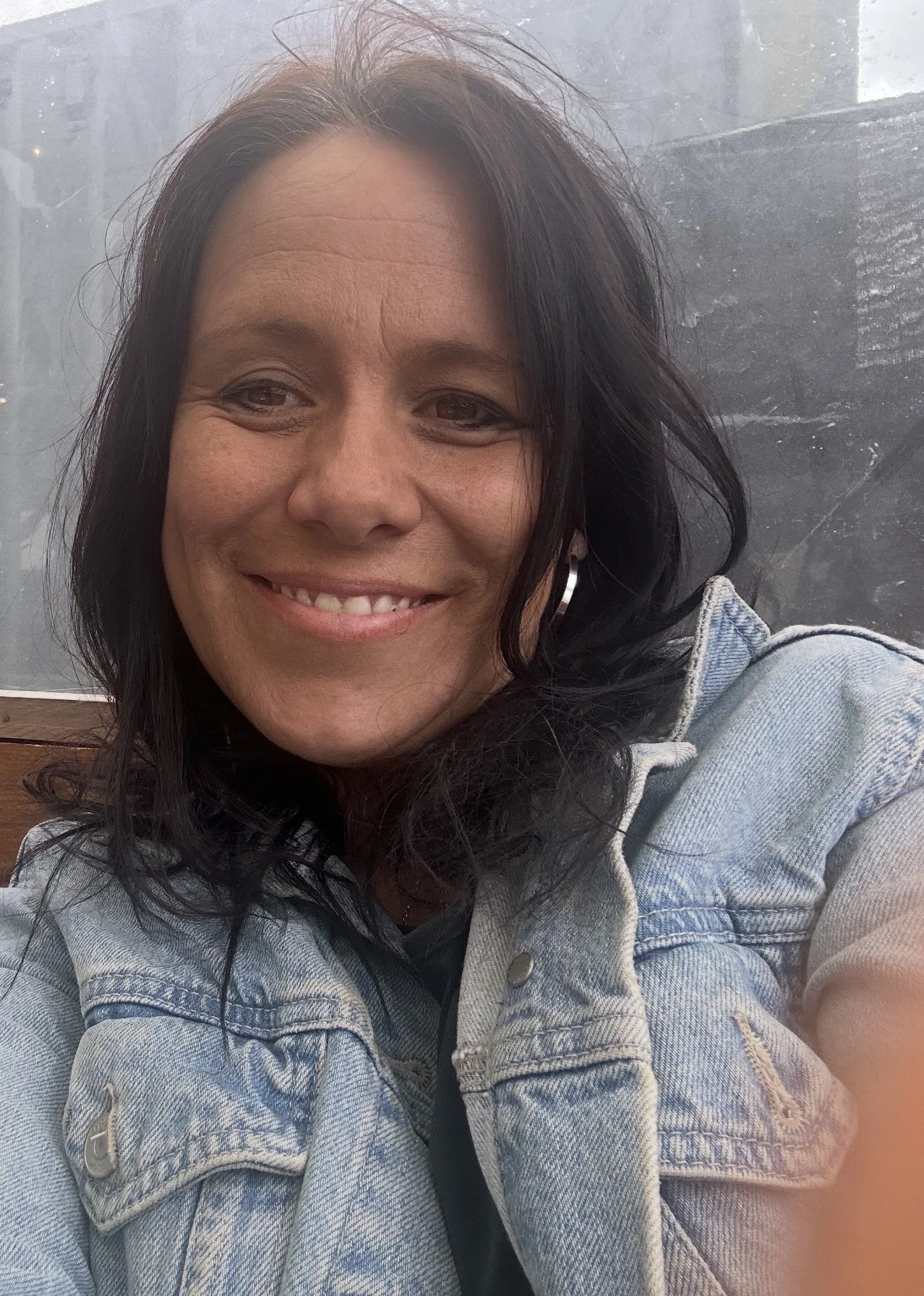 A woman with dark wavy hair smiling at the camera, wearing a light denim jacket, sitting in front of a window with a cityscape in the background.