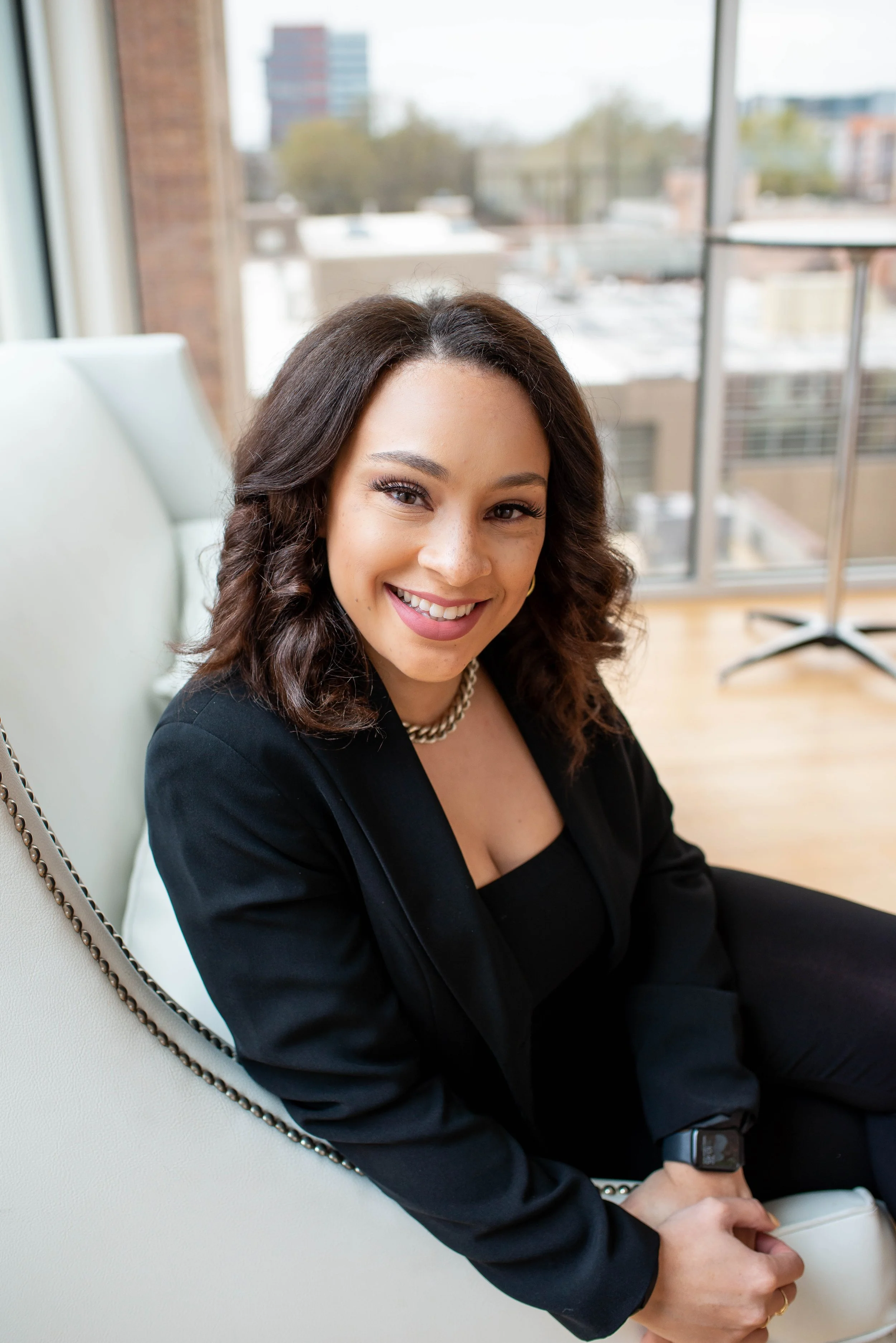 A woman with shoulder-length wavy hair, wearing a black blazer, sitting on a white armchair, smiling at the camera with a city view visible through large windows behind her.
