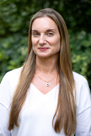A woman with long brown hair and blue eyes standing outdoors with green foliage in the background, wearing a white top and a gold necklace with a blue pendant.