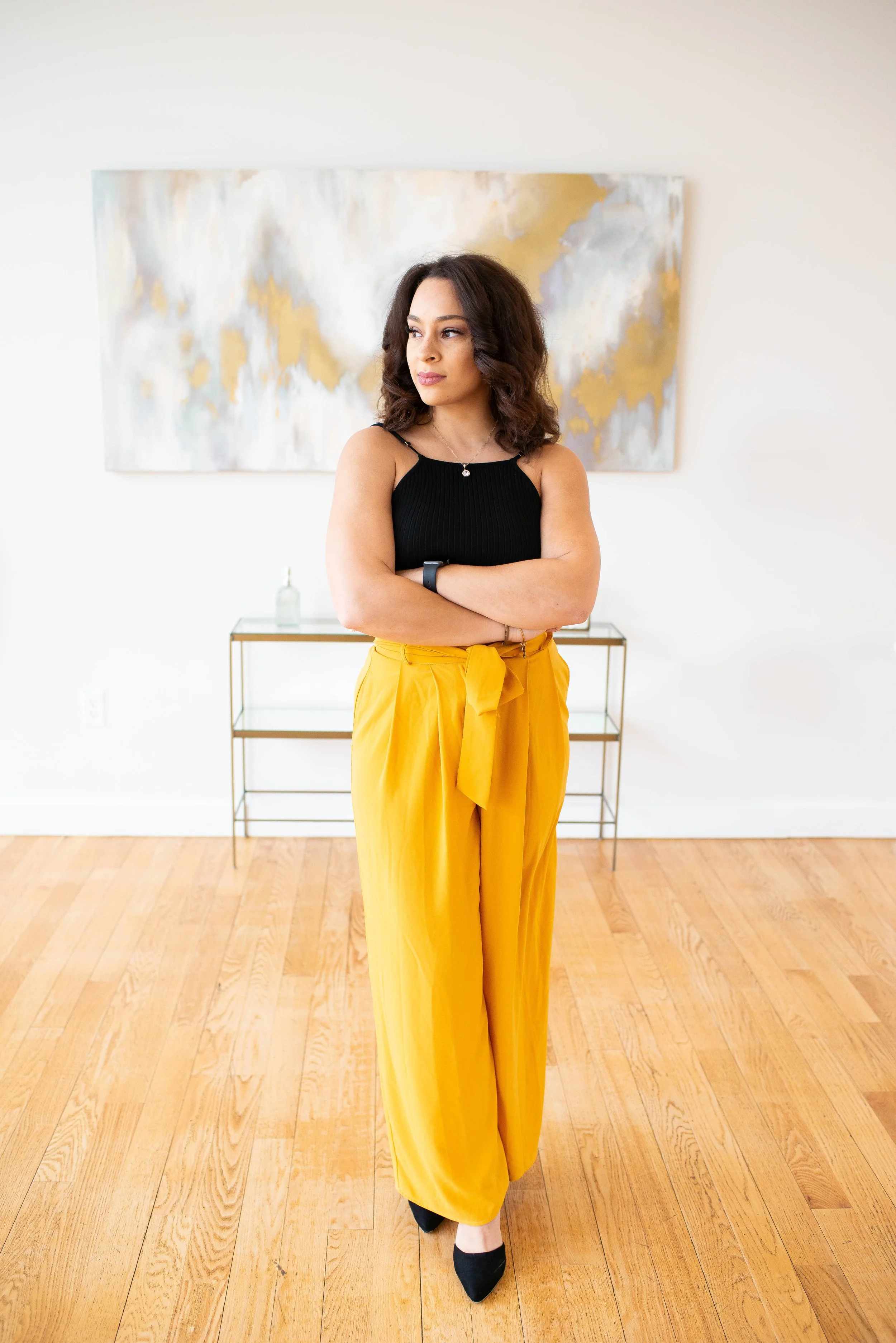 A woman with wavy dark hair, wearing a black sleeveless top, yellow high-waisted pants with a bow, black heels, standing with arms crossed in a bright room with wooden floors and modern art on the wall behind her.