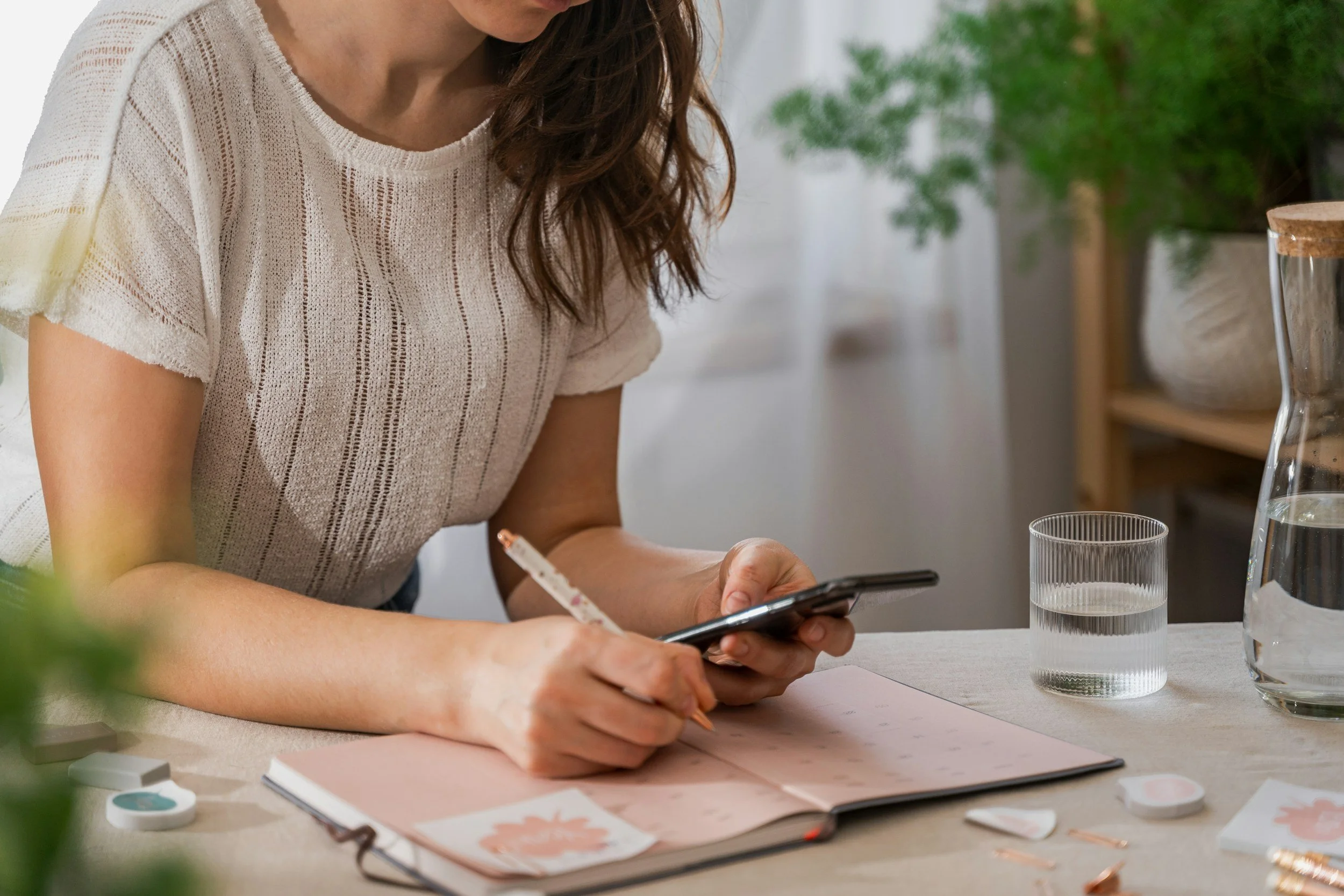 A woman is sitting at a table, writing in a planner or journal while looking at her phone, with a glass of water and a pitcher on the table. There are plants and home decor in the background.