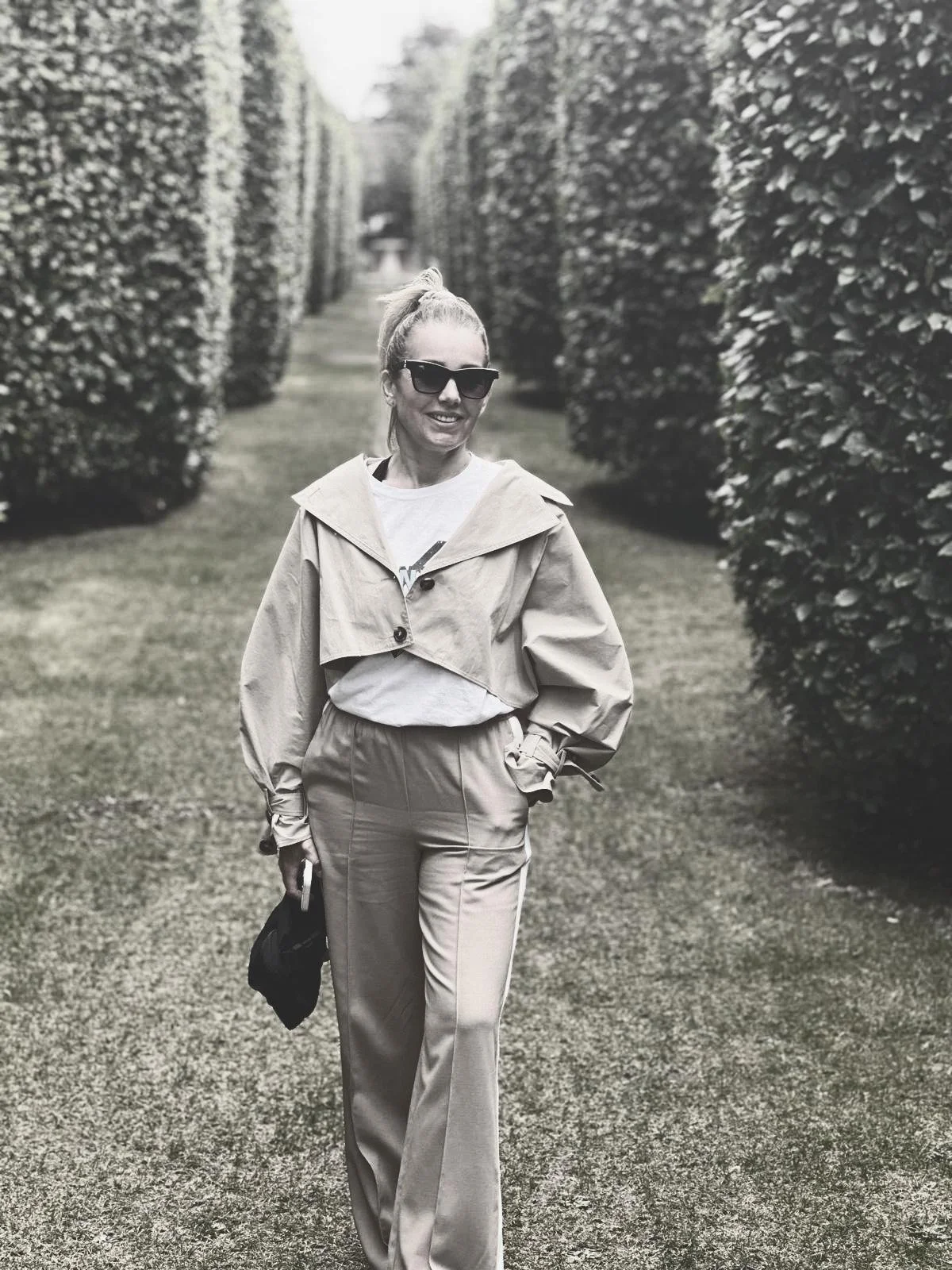 A woman walking along a path with tall, neatly trimmed hedges on both sides, wearing sunglasses, a light-colored jacket, casual pants, and holding a small bag and a phone.