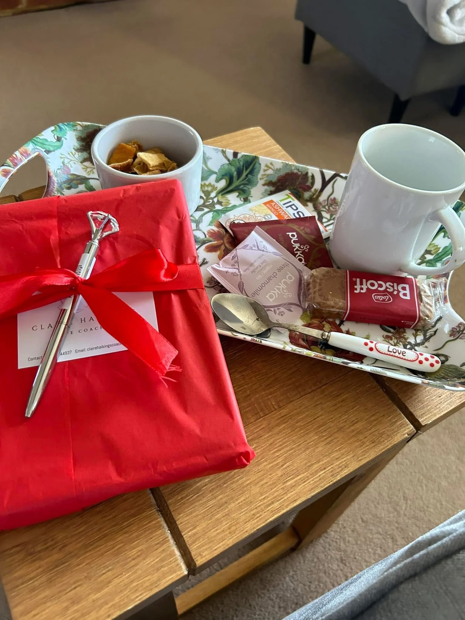 A tray with tea or coffee, biscuits, and tea bags on a wooden table, next to a wrapped gift with a toy razor, in a cozy room.