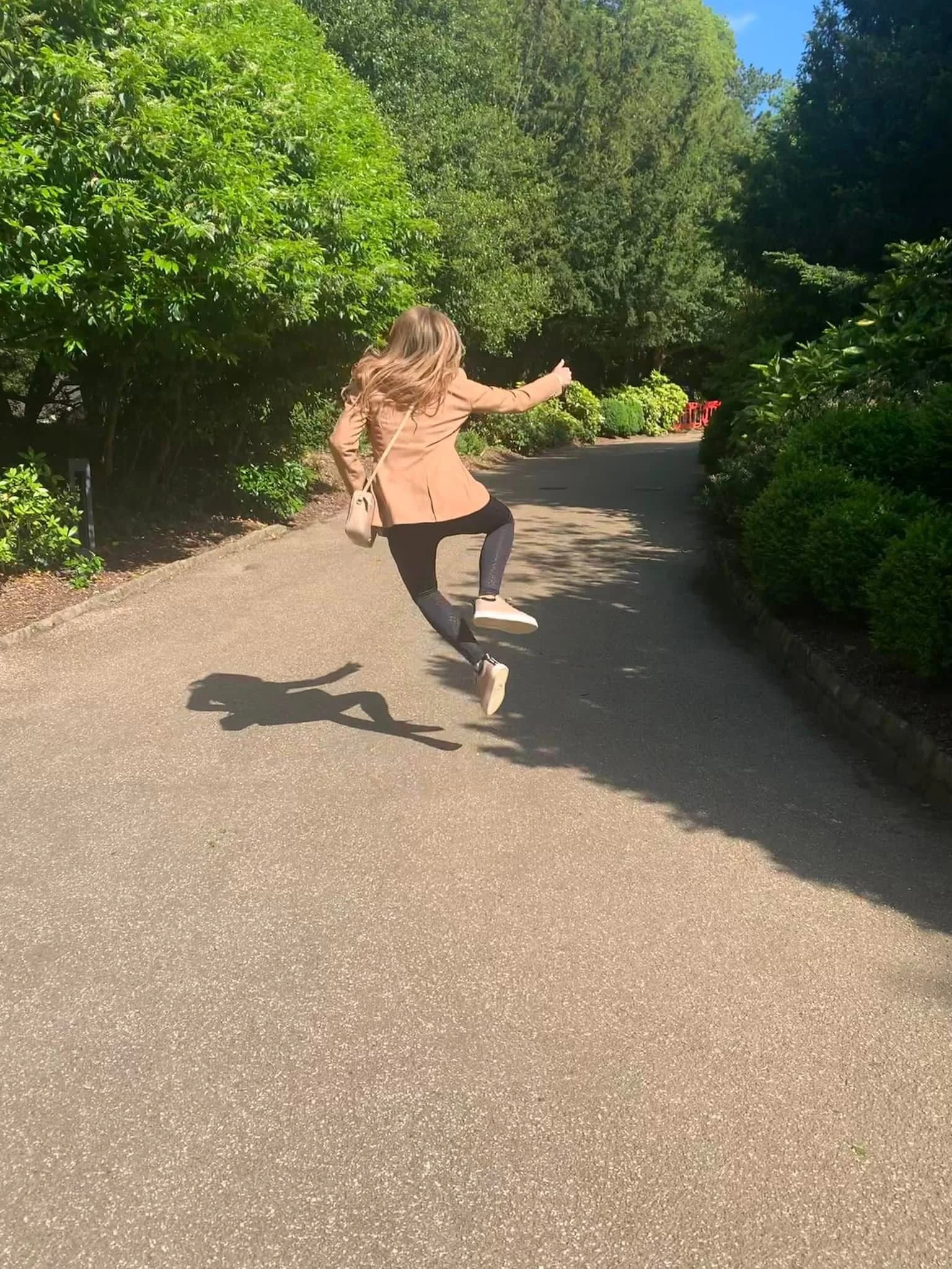 A woman with long hair jumping in the air on a paved pathway surrounded by greenery and bushes on a sunny day.