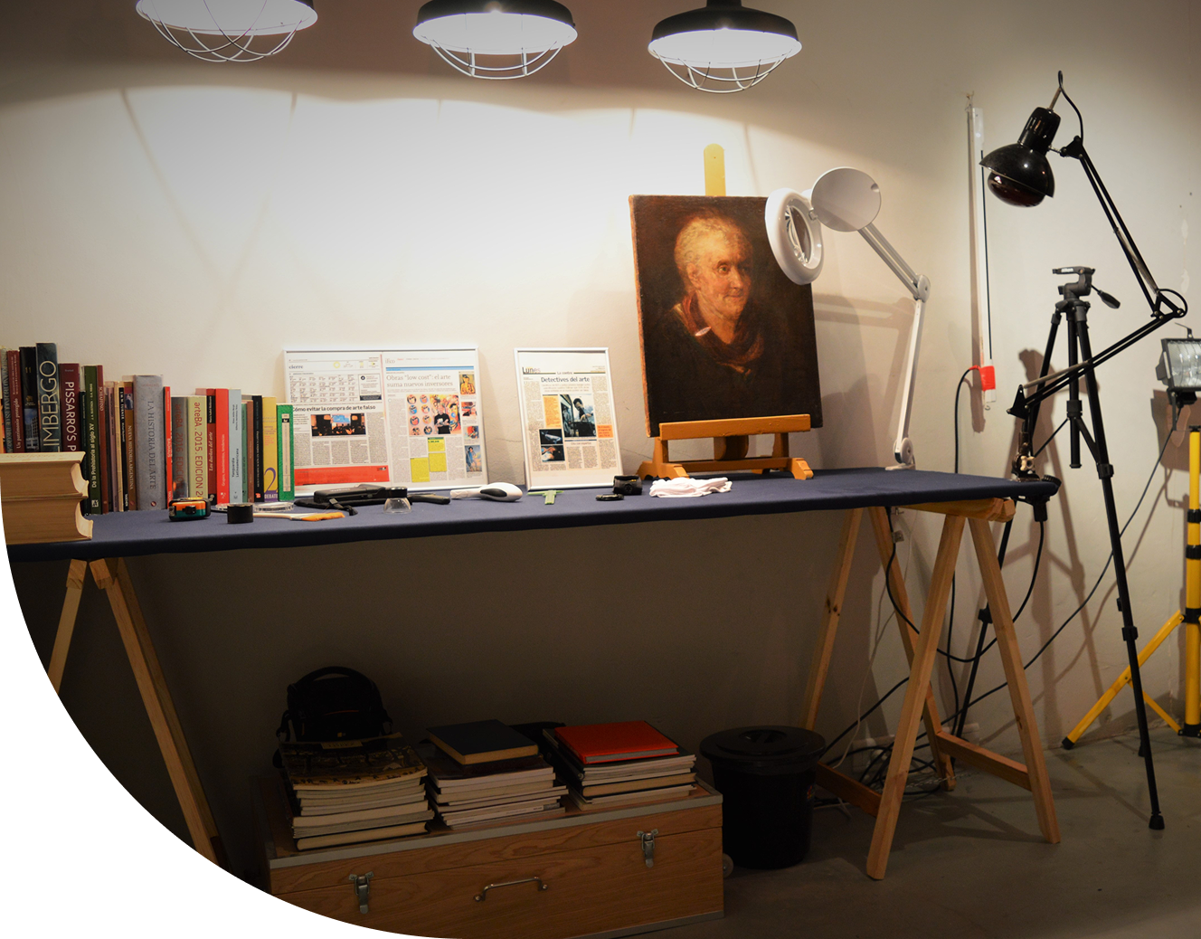 Estúdio de arte com uma mesa de trabalho, livros, uma pintura de retrato e equipamentos fotográficos.