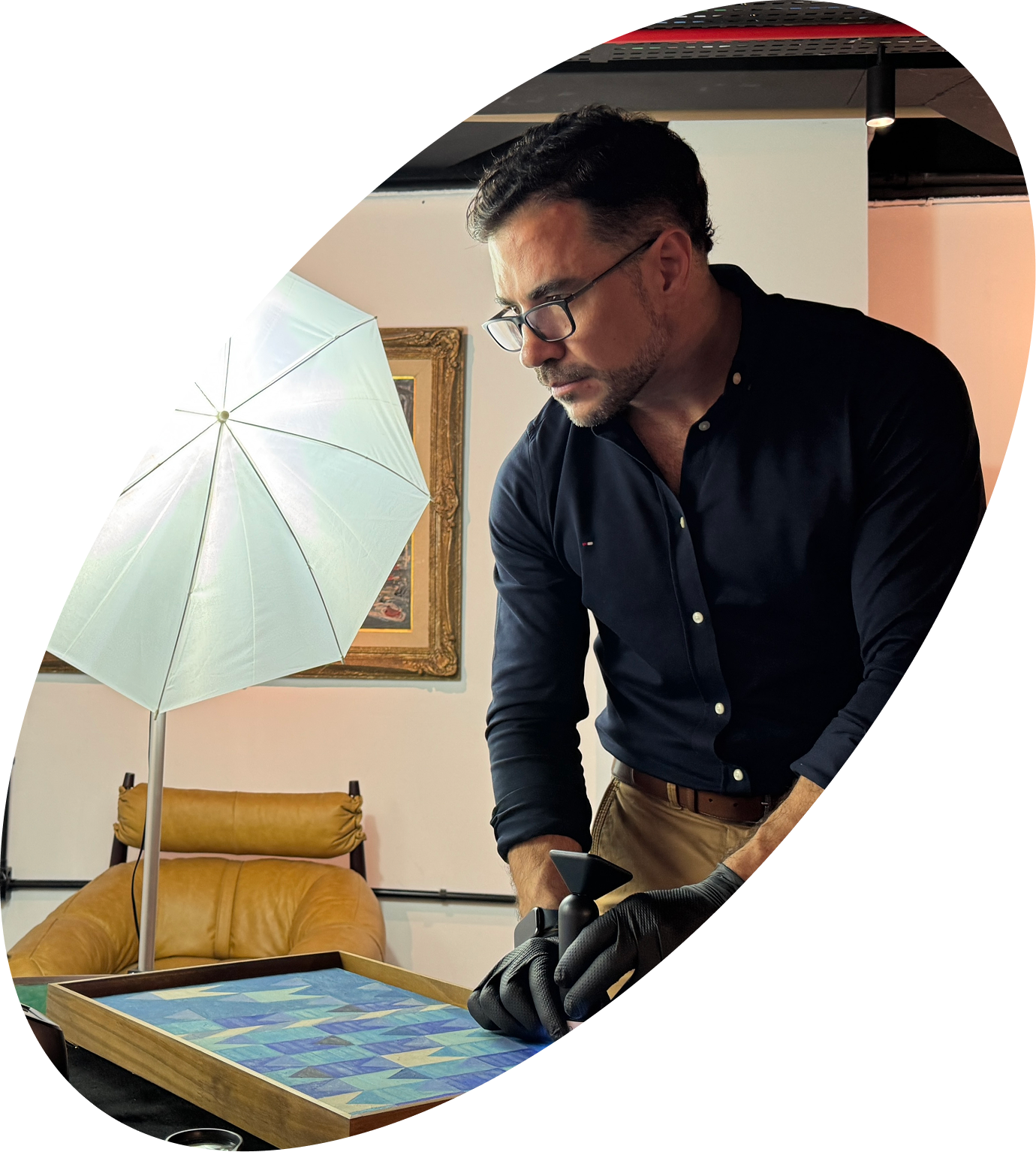 Homem com óculos de grau e camisa azul escura, usando luvas pretas, manuseando uma obra de arte em uma mesa de madeira, em um ambiente com quadro na parede e uma cadeira de couro marrom ao fundo.