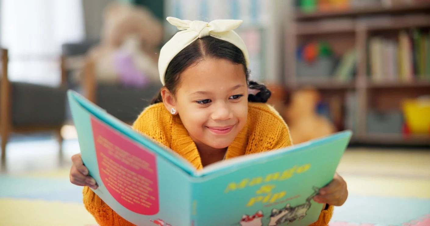Young girl in a yellow sweater and white headband reading with more confidence because of reading tutoring in St. Charles, MO, with a bookshelf and a dog in the background.