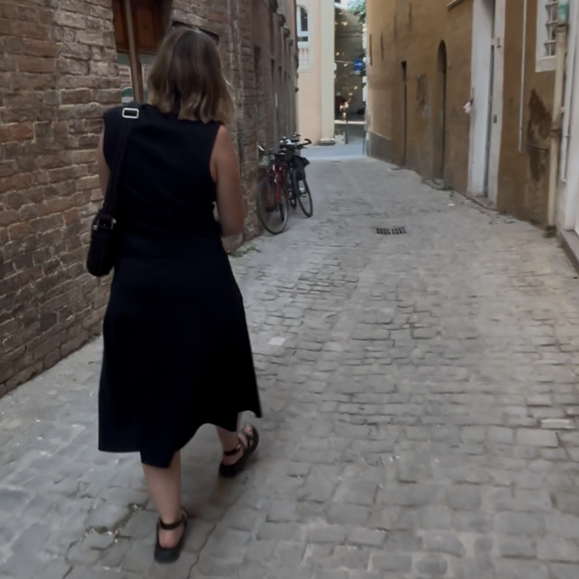 Poster for Guided Tour of the Untrue History. Woman walking down a narrow cobblestone alleyway with bicycles parked against the brick wall on the left and buildings on the right.