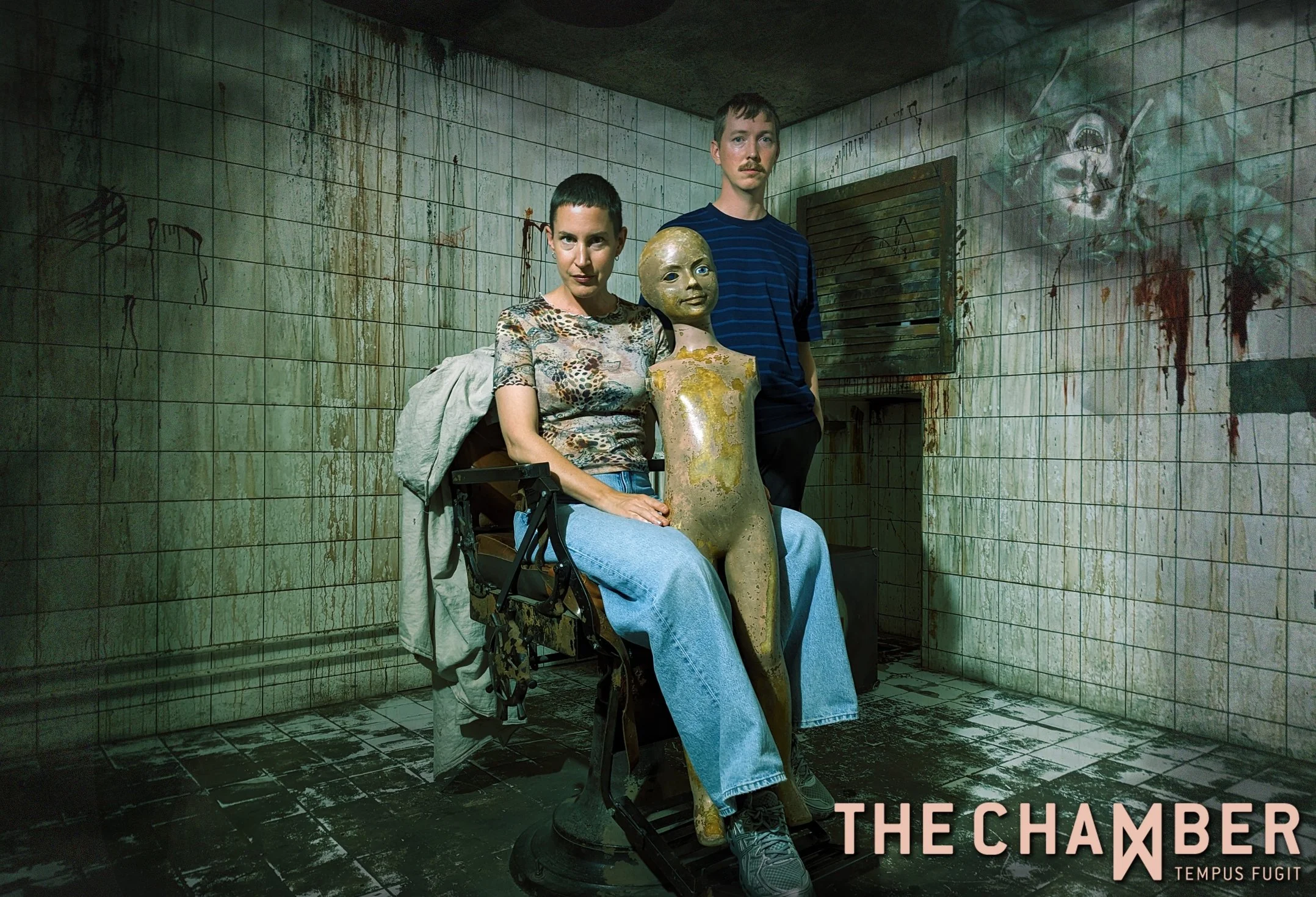 Poster for The Escaperoom. Two people, a woman and a man, sit and stand next to a distorted doll in a grimy, dirty room with dirty tiled walls and floors, and graffiti on the walls.