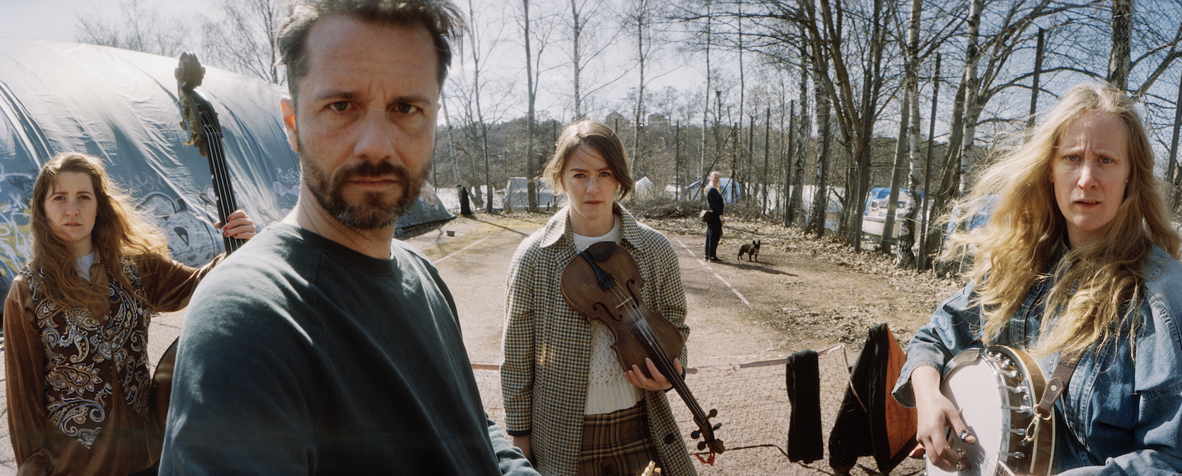 Picture from Fight/ Bråk. Four people outdoors, two women and two men. The man in front has a beard, the woman in the middle holds a violin, and the woman on the right plays a tambourine. A person in the background has an acoustic bass.