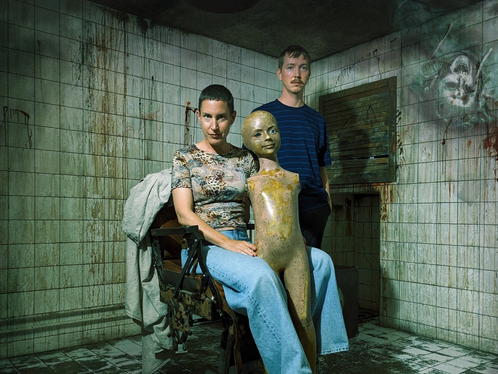 Poster for The Escaperoom. Two people, a woman and a man, sit and stand next to a distorted doll in a grimy, dirty room with dirty tiled walls and floors, and graffiti on the walls.