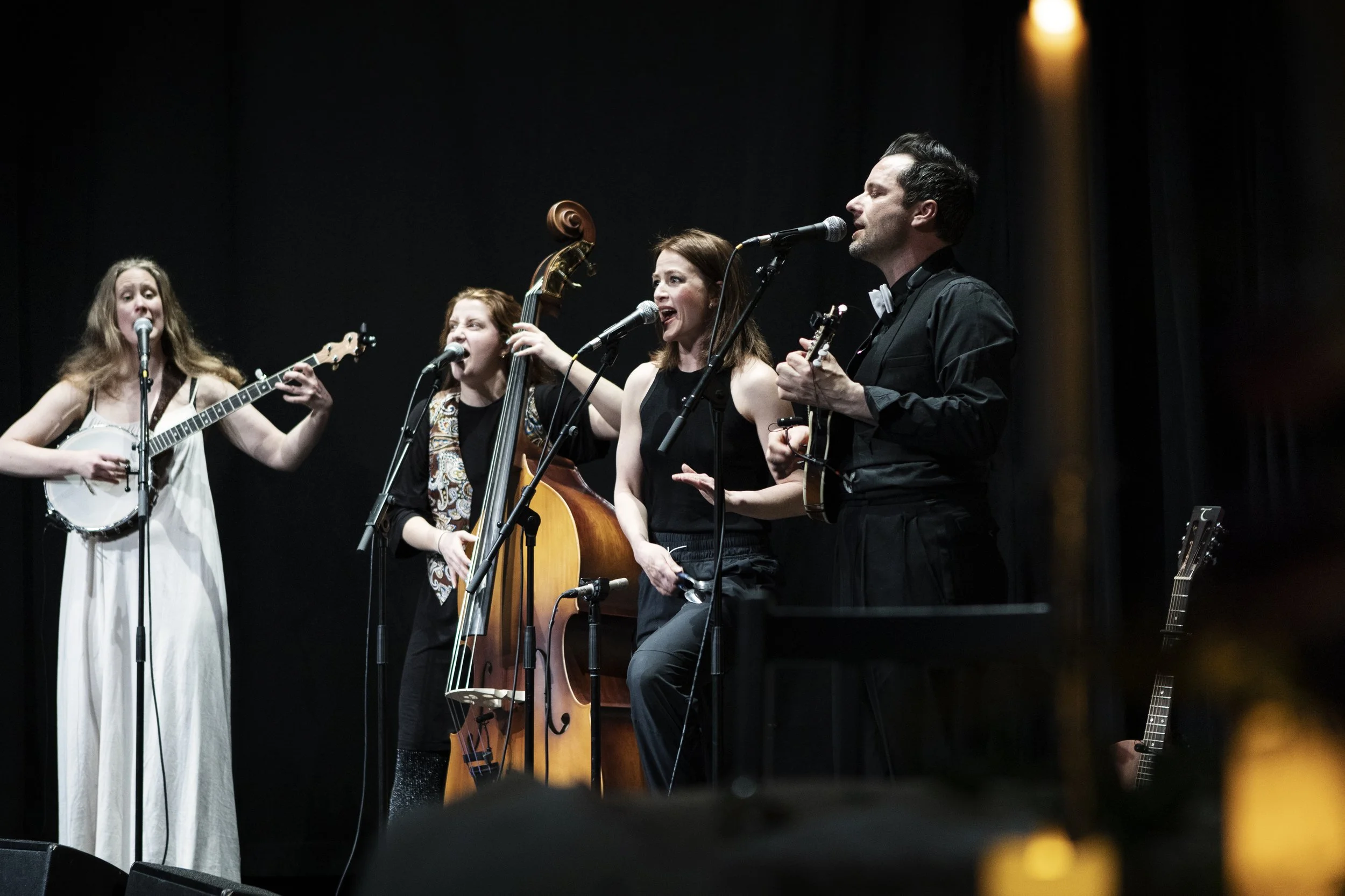 Picture from Fight / Bråk. Four musicians performing on stage: a woman with long hair playing a banjo, another woman with red hair playing a double bass, a woman with short hair singing, and a man with dark hair playing a ukulele.