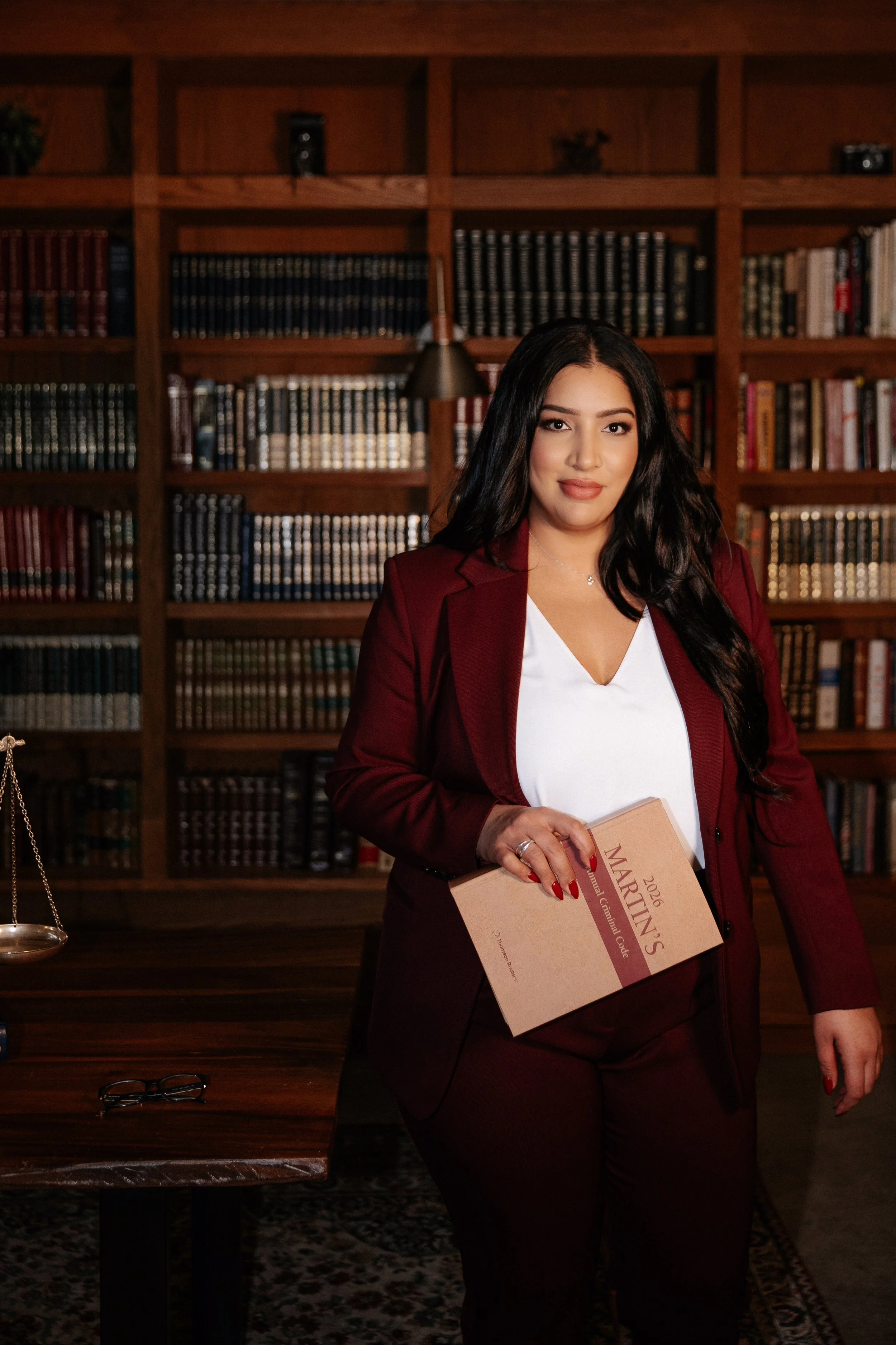 A professional woman with long dark hair standing in a library holding a book titled 'Martins 2020 Criminal Code'. She wears a burgundy suit with a white blouse, and there are shelves of books and a scale on a desk in the background.
