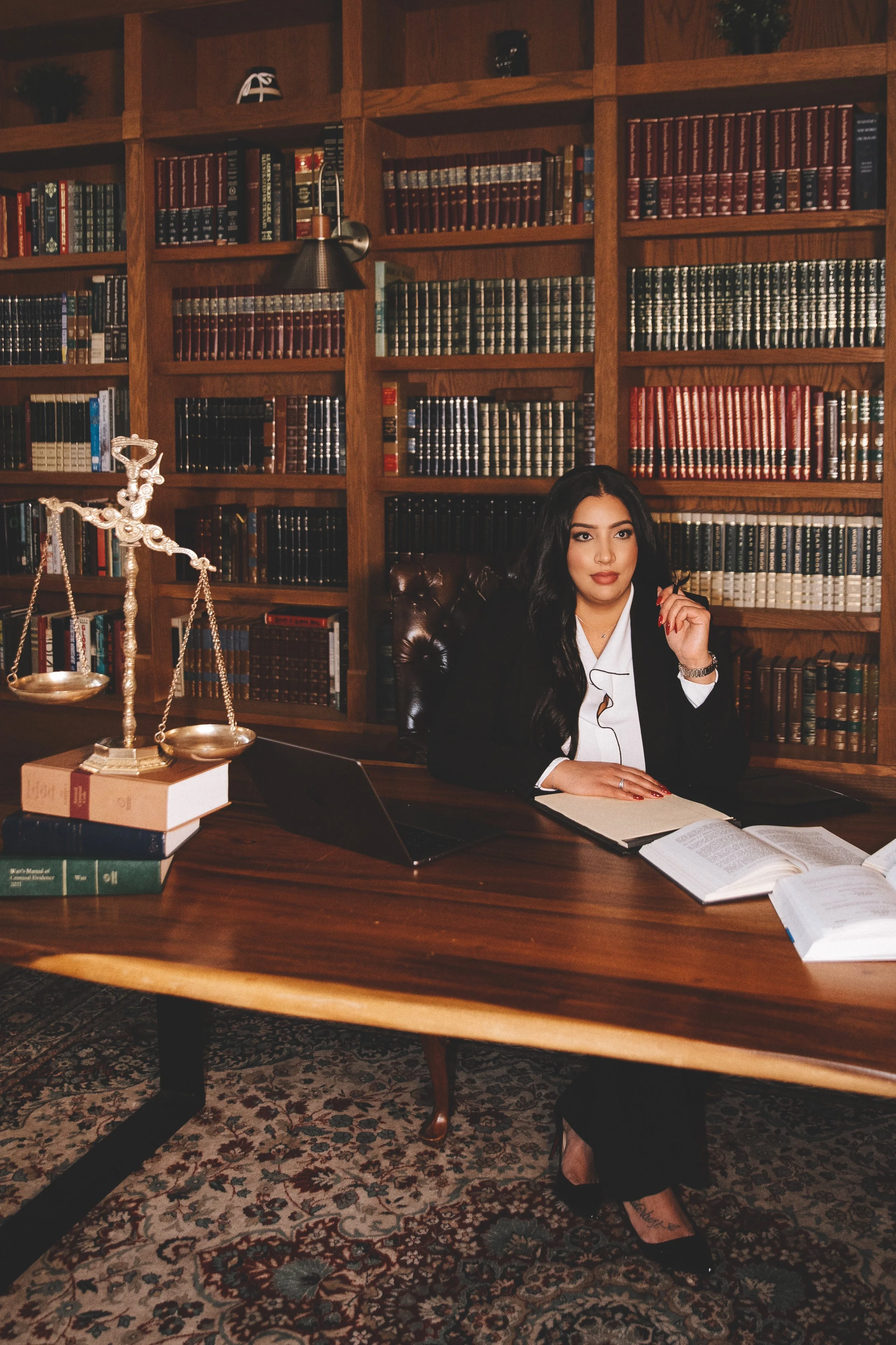 A woman sitting at a wooden desk in a library, surrounded by bookshelves filled with books, with legal books, laptop, and legal scales on the desk.