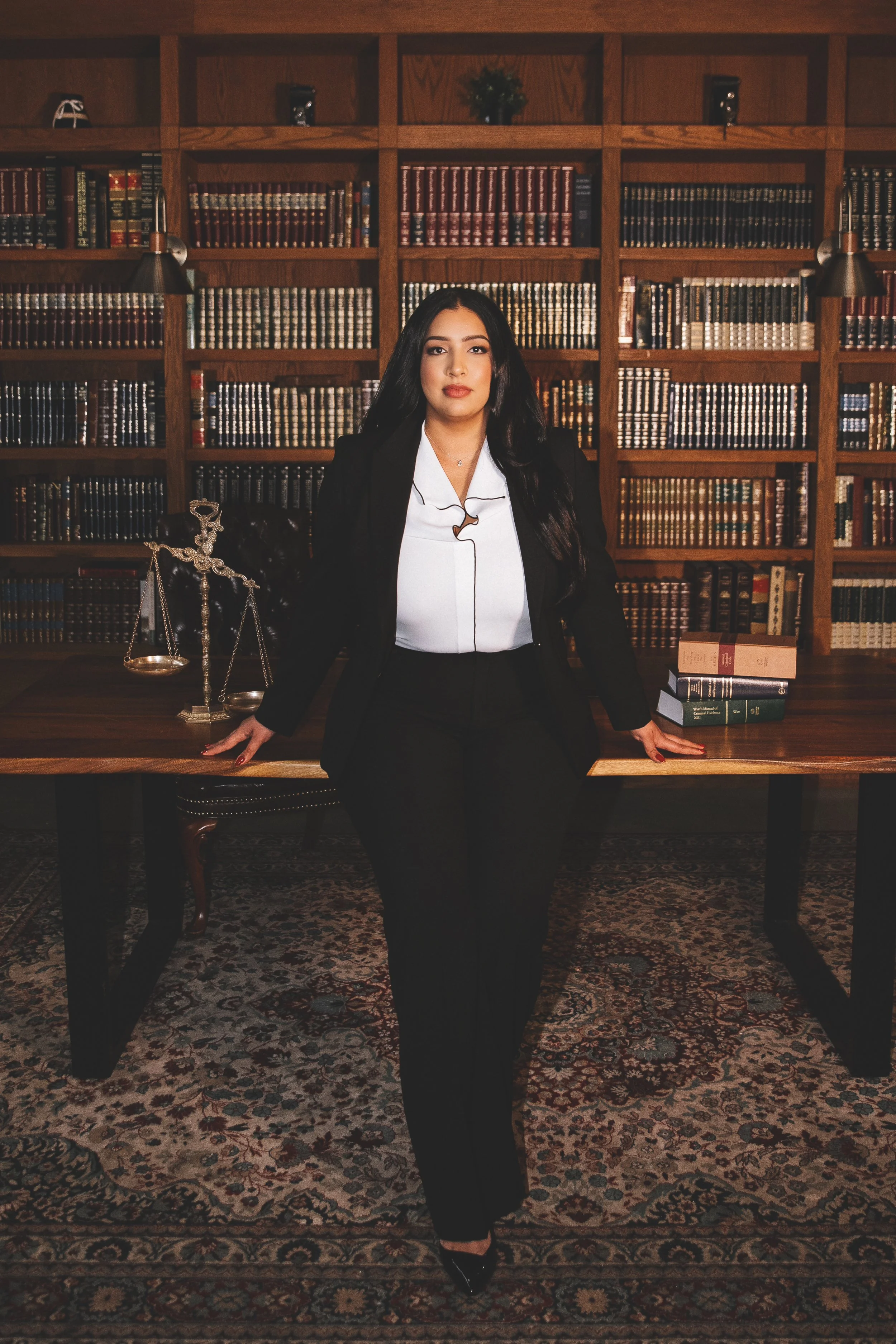 A woman in a black suit and white blouse standing in a library, with a wooden table, legal scales, and stacks of books behind her.