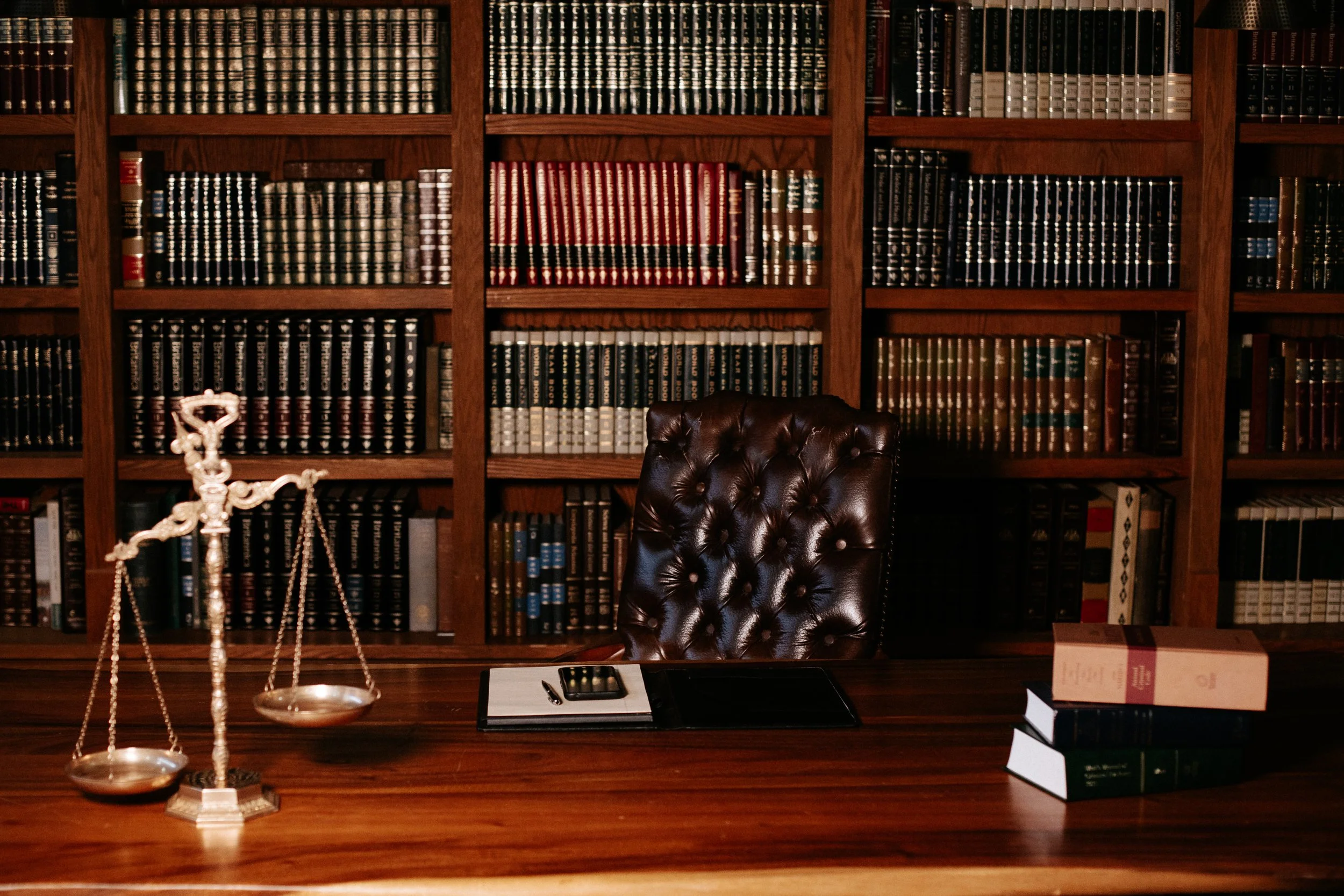 A professional office with a large wooden desk, a leather chair, a set of bookshelves filled with legal books, a brass balance scale, a notepad, a pen, a phone, and three stacked legal books.