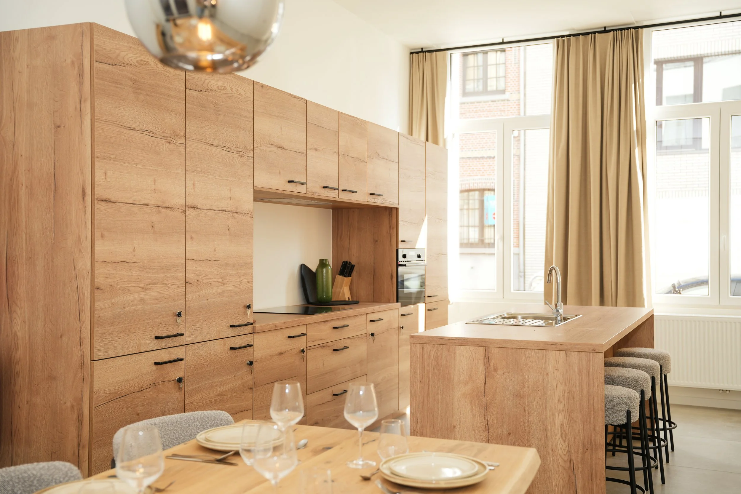 Een moderne keuken met houten kasten en een eiland, grote ramen met beige gordijnen en een eettafel met servies en glazen.