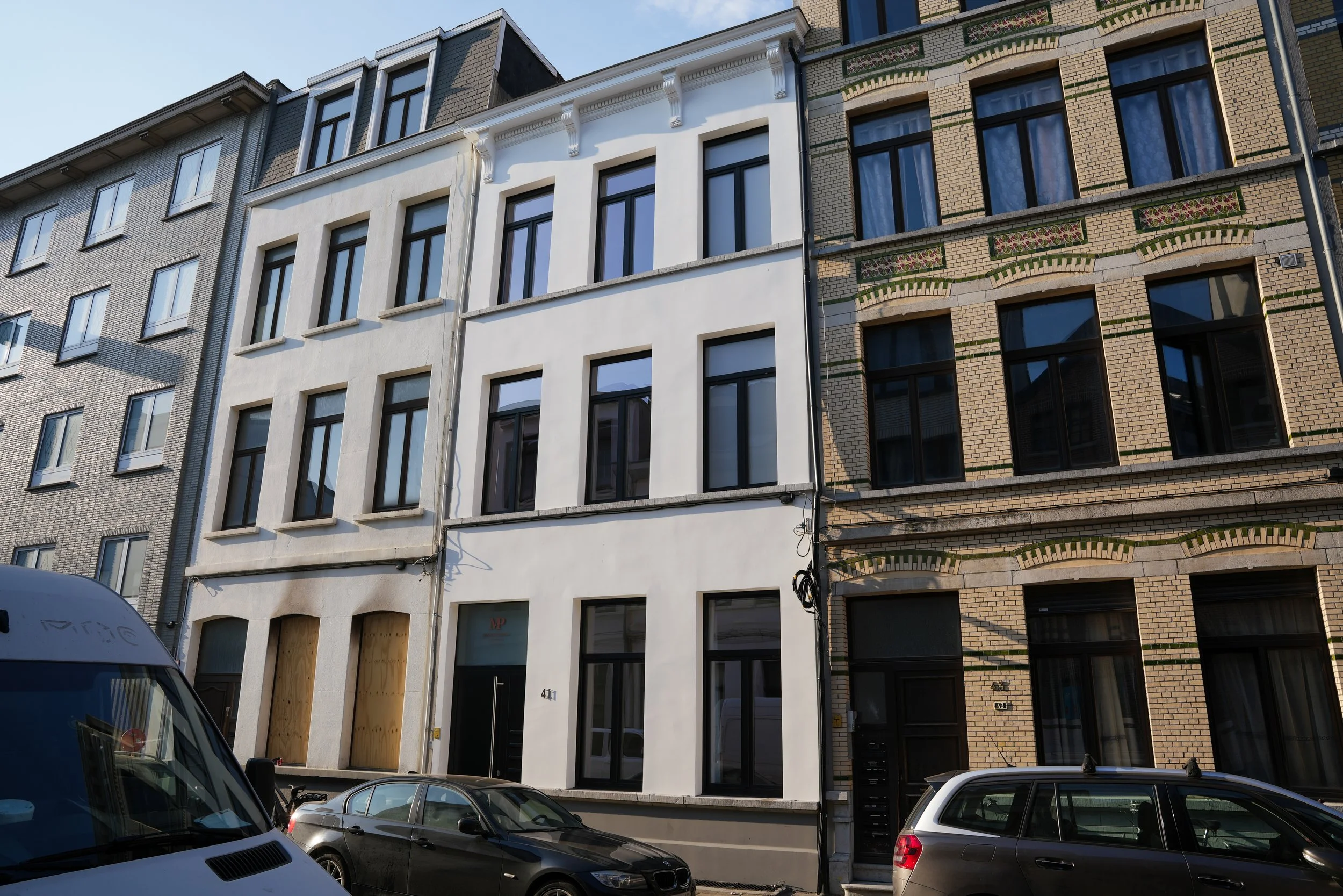 Drie gebouwen met verschillende gevels en ramen, met auto's geparkeerd op de straat voor de gebouwen.