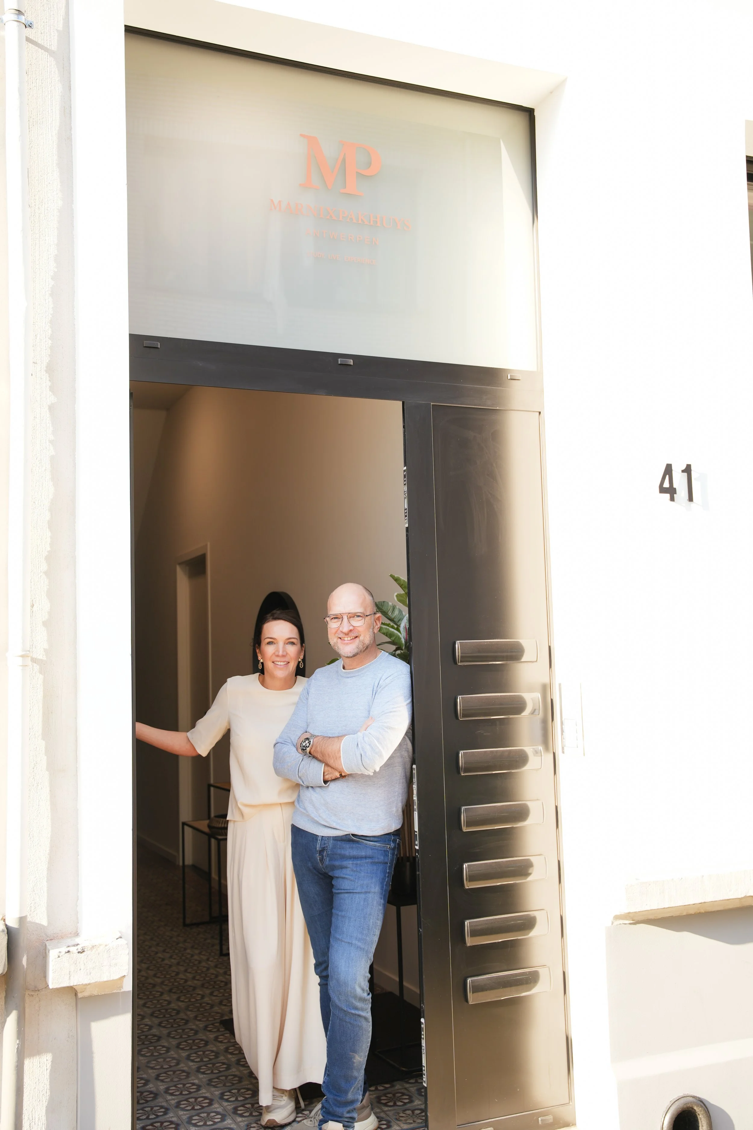 Twee mensen, een man en een vrouw, staan bij de ingang van een woning, glimlachend naar de camera. De man draagt een grijs shirt en blauwe jeans, de vrouw een witte jurk. De voordeur is open, binnen is een gang met een tafel en decoratie te zien. Nummer 41 is zichtbaar op de muur naast de deur. Boven de deur hangt een glazen plaat met de tekst 'MARNIXPAKHUYS ANTWERPEN'.