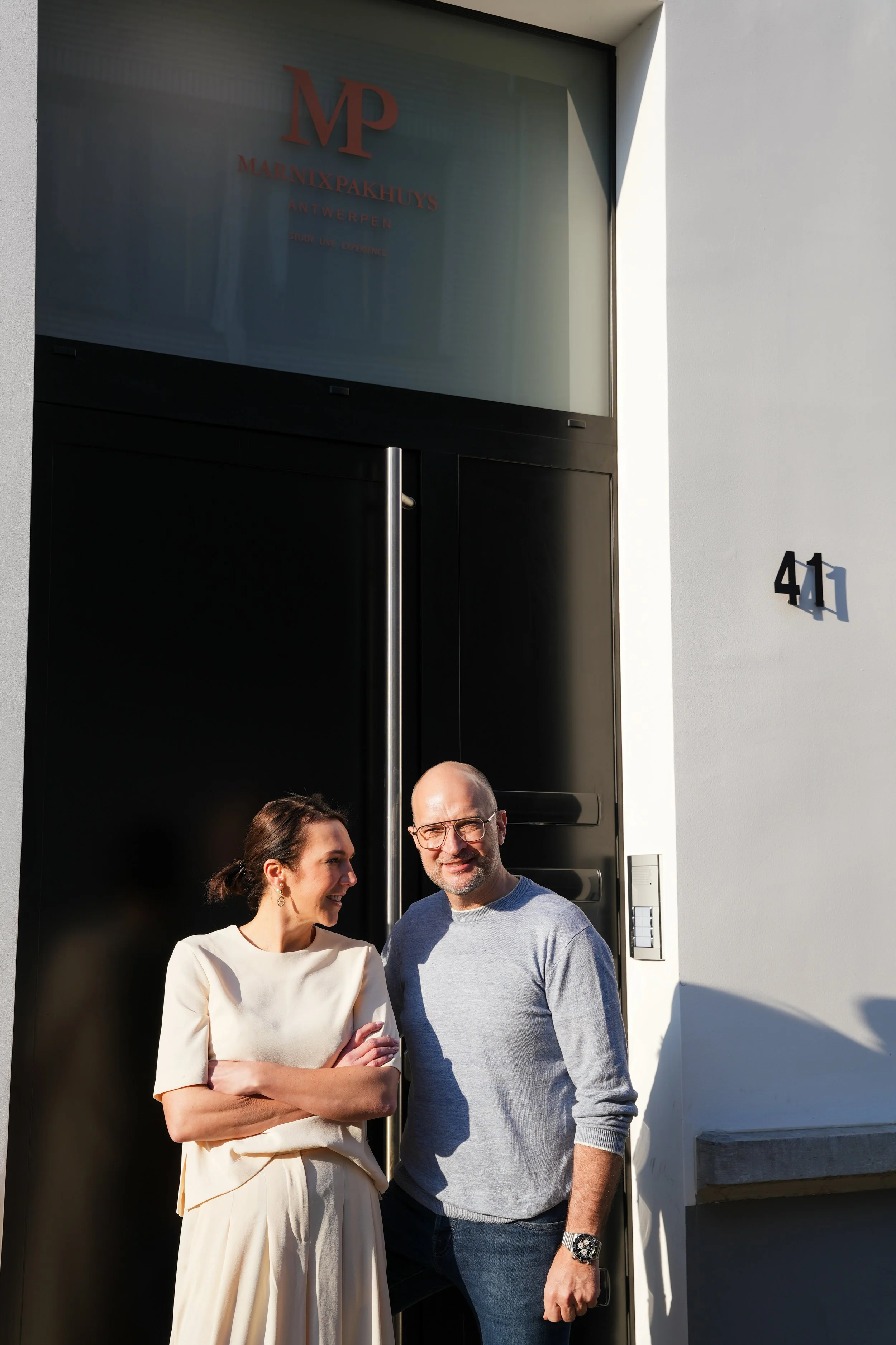 Twee mensen, een vrouw en een man, staan buiten voor een zwart deur met nummer 41, lachend en pratend, met een wit gebouw op de achtergrond.