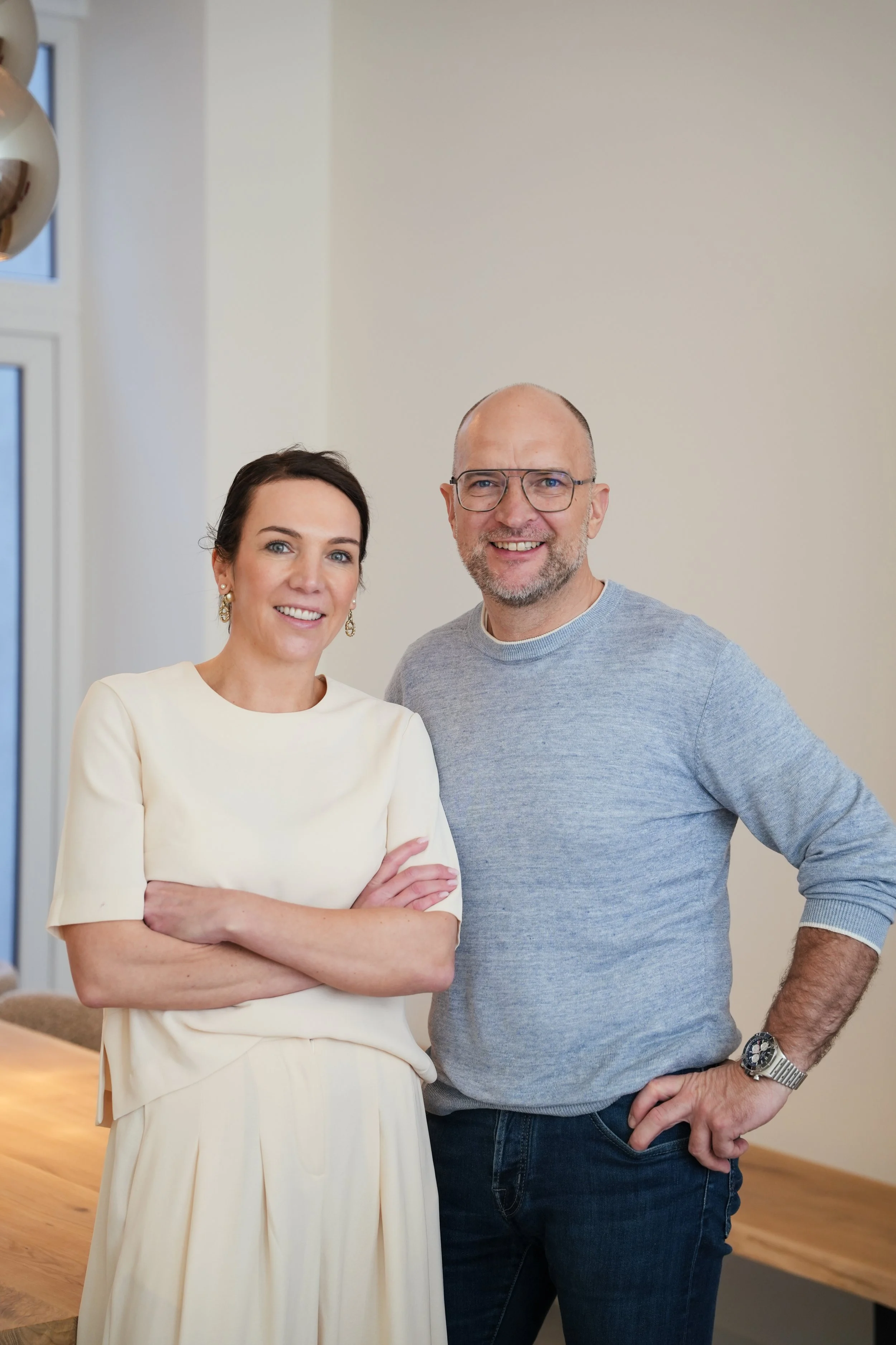 Een man en een vrouw staan samen binnen, beiden glimlachend naar de camera. De vrouw draagt een witte jurk met korte mouwen en heeft donker haar, de man draagt een grijs trui en bril, en heeft een baard en een gladgemaakt hoofd. Ze lijken gelukkig en ontspannen.