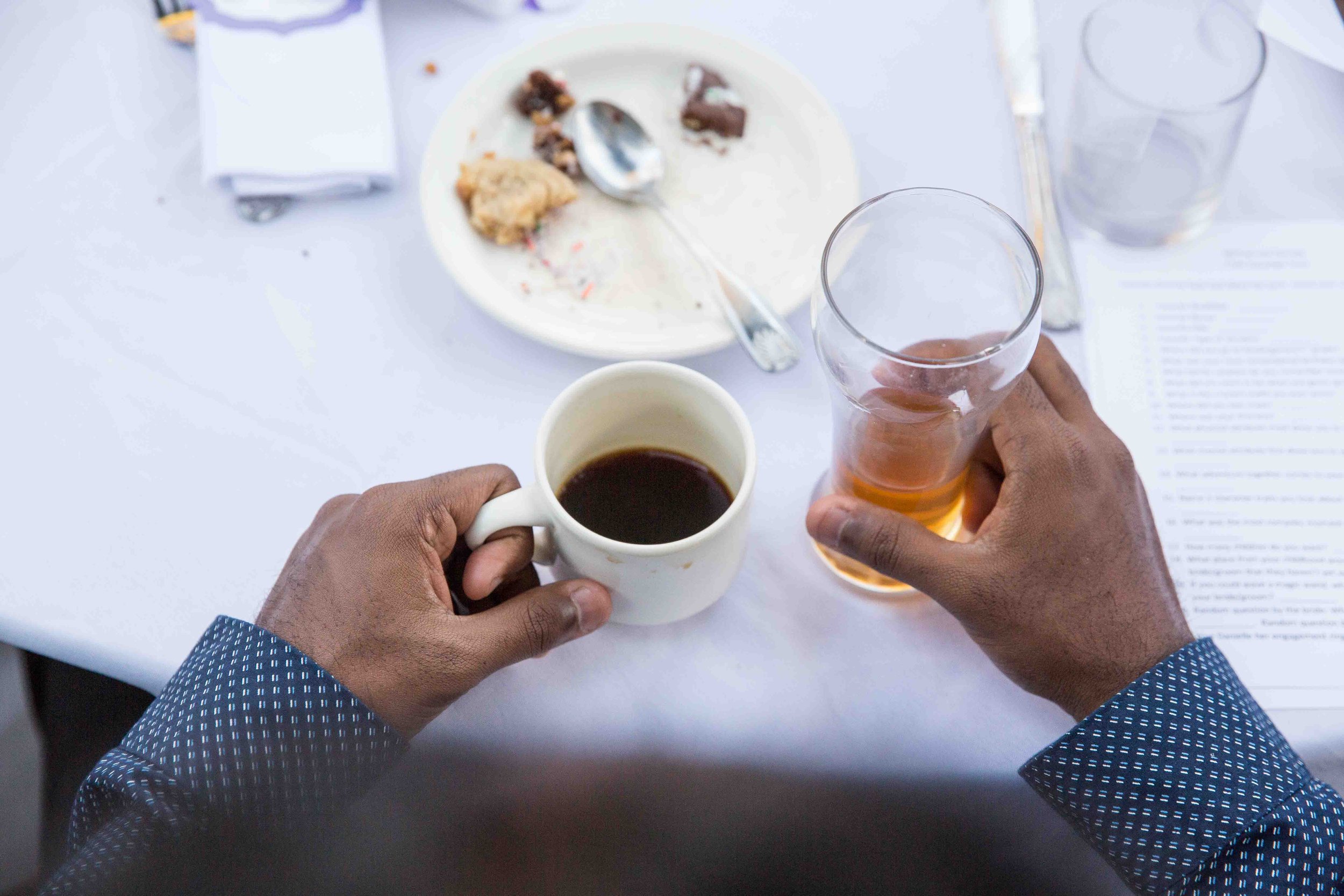 Person holding a white coffee mug and a glass of orange-colored beverage over a white table with a white plate containing leftover dessert, a spoon, a glass of water, and a paper menu.