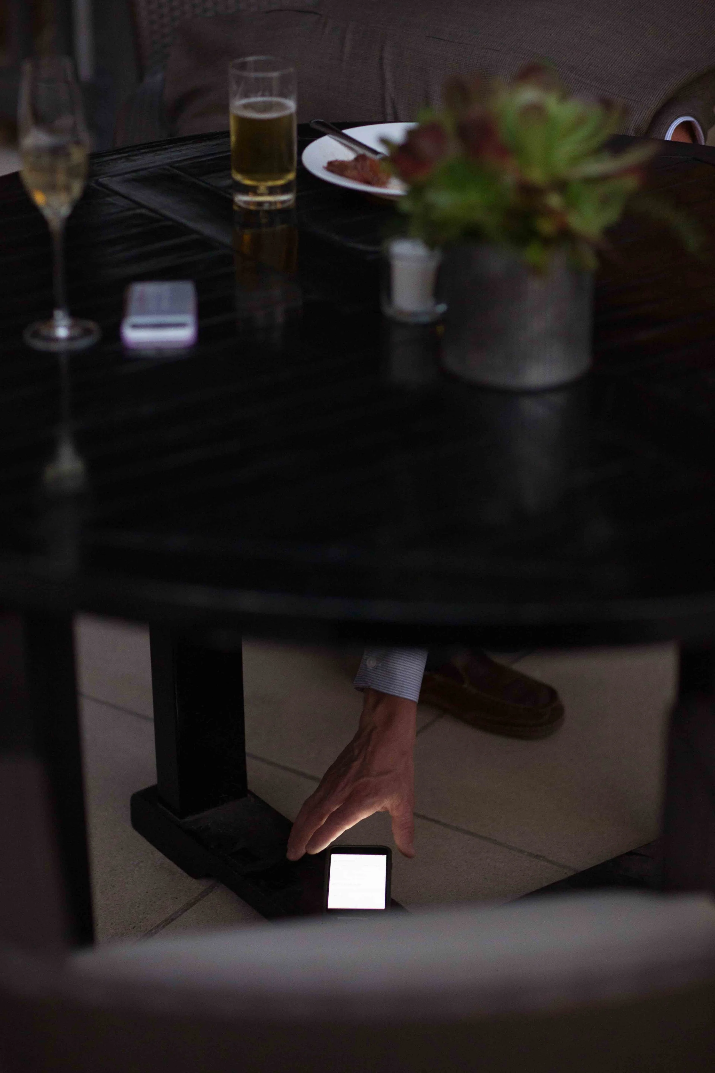 The image shows a person sitting at a dark wooden table with drinks and a plate of food. Below the table, a hand is seen reaching to adjust a small device on the floor, which has a bright screen.