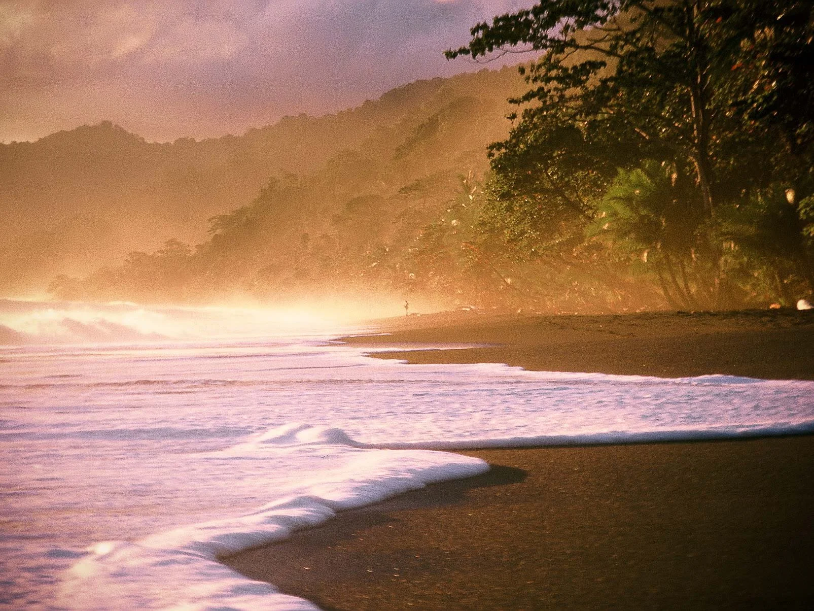 Sunset over a tropical beach with dark sand, gentle waves, and lush green trees on a hillside.