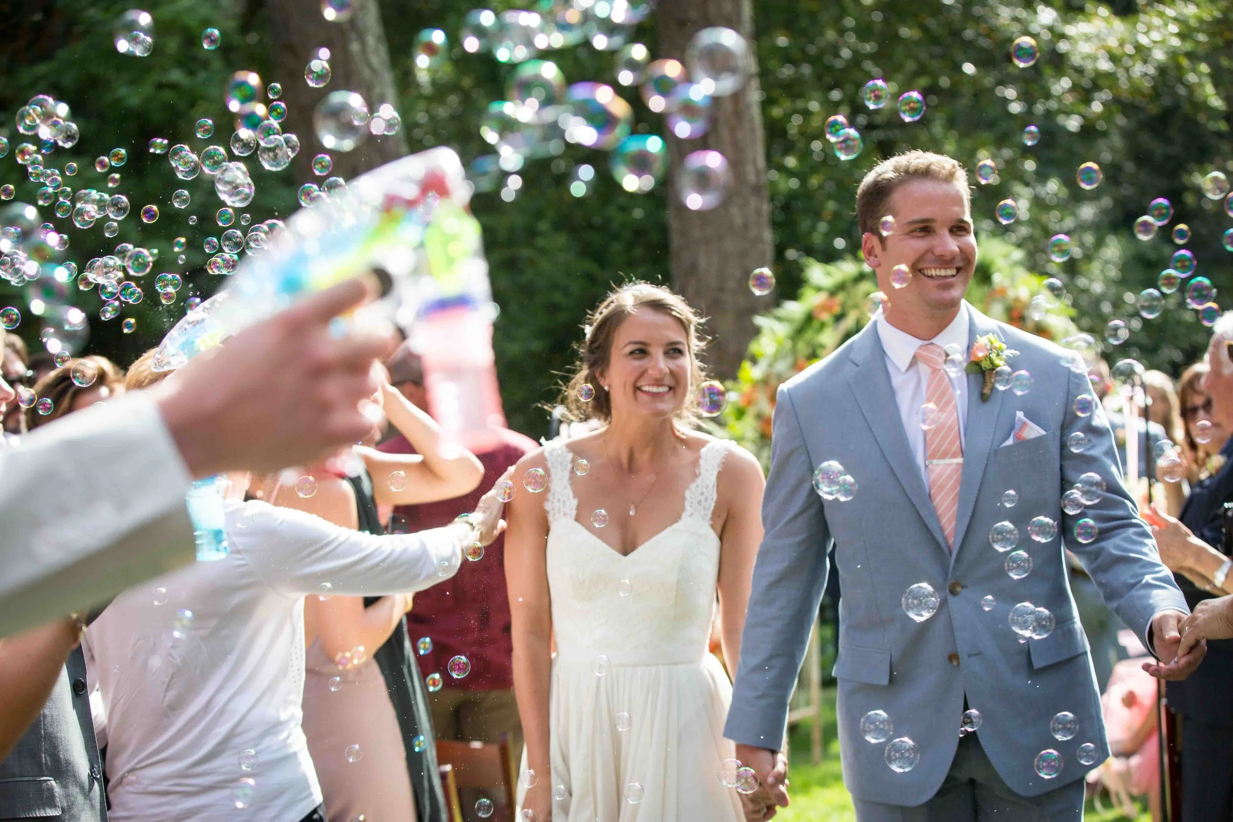 A newlywed couple walking hand in hand at an outdoor wedding celebration, surrounded by guests and bubbles in the air, with a lush green background.